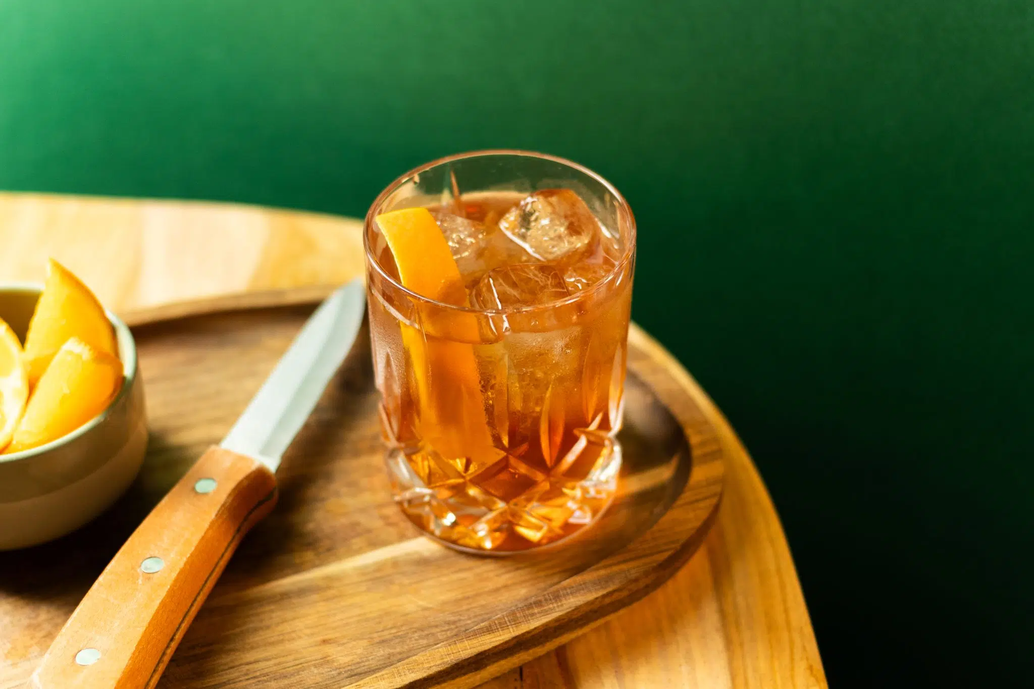 A high angle shot of a Maple Old Fashioned cocktail in an old fashioned glass on a wooden board on a wooddedn table with a knife and a bowl with orange wedges around, in front of a green background.