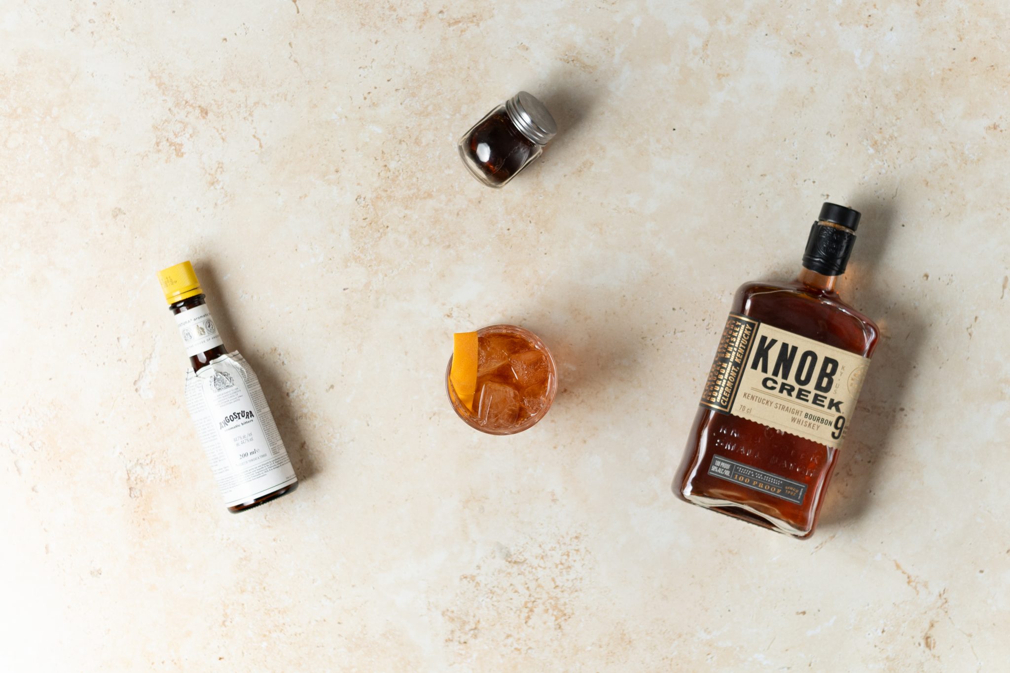 A Maple Old Fashioned cocktail, shot from above, in an old fashioned glass on a beige surface surrounded by Bourbon, maple syrup and Angostura aromatic bitters