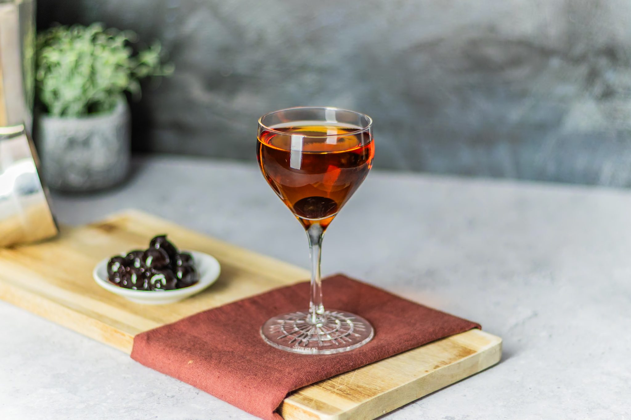A side shot of a Martinez cocktail in a coupe glass on a maroon cloth with a plate full with Maraschino cherries to the side on a wooden board placed on a white table in front of a grey stone wall.