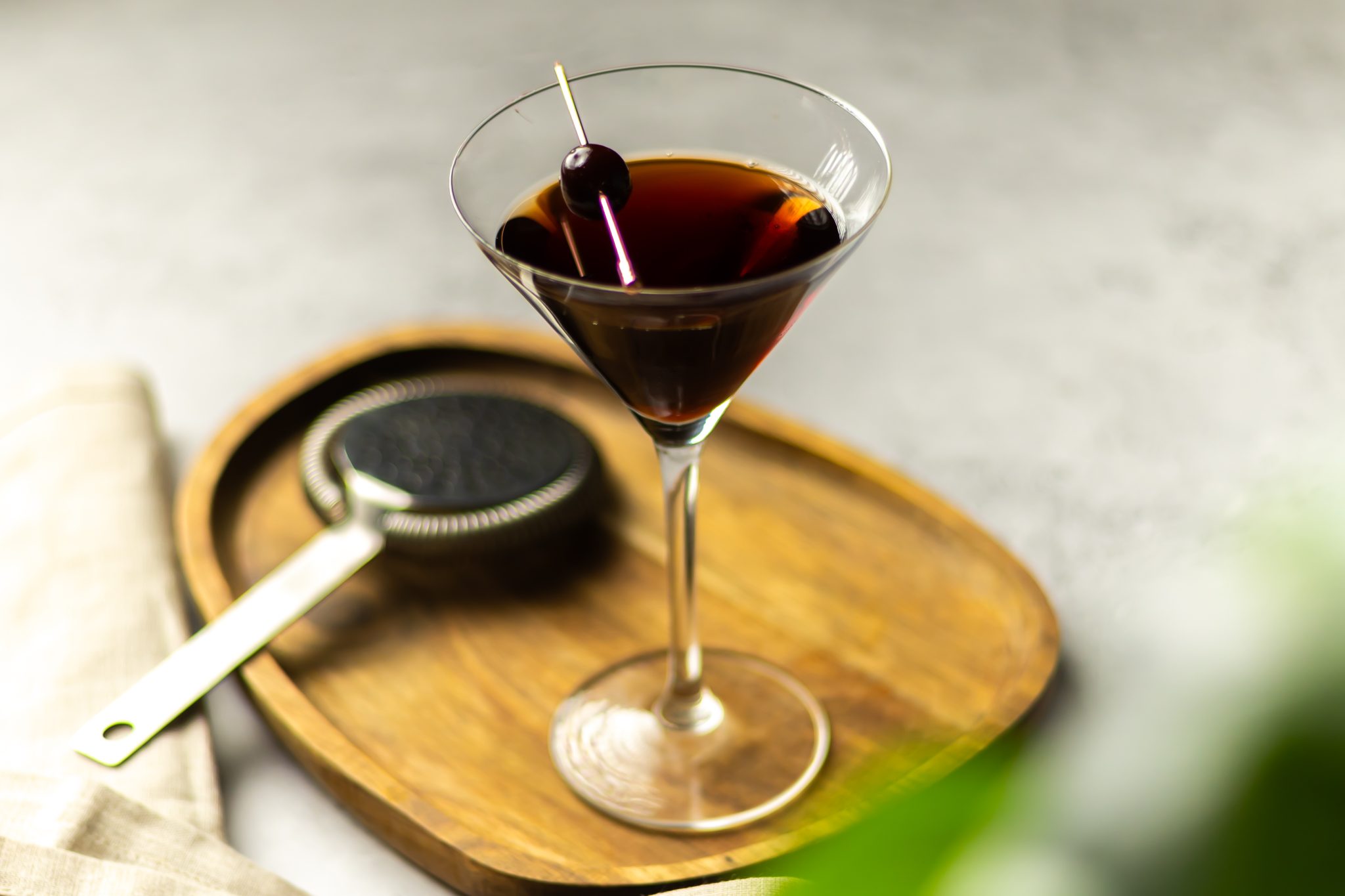 A side shot of a Metropolitan cocktail in a martini glass on a wooden tray placed on a grey table surrounded by a strainer, and a bar spoon