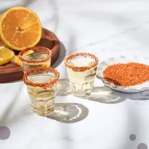 Three shots glasses with spices over the rim filled with mezcal spirit, a plate with spices and a wooden board with orange pieces on top, placed on a white table.
