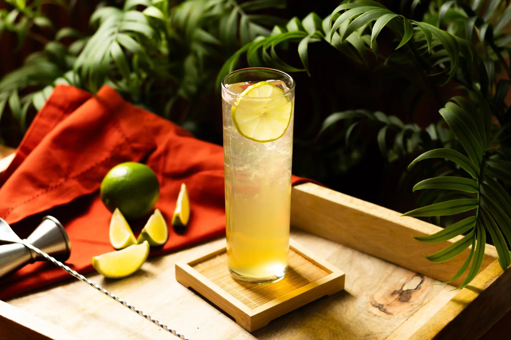 A side shot of a Mezcal Collins cocktail in a collins glass on a wooden coaster and tray with four lime wedges, a lime, a red cloth and a bar spoon