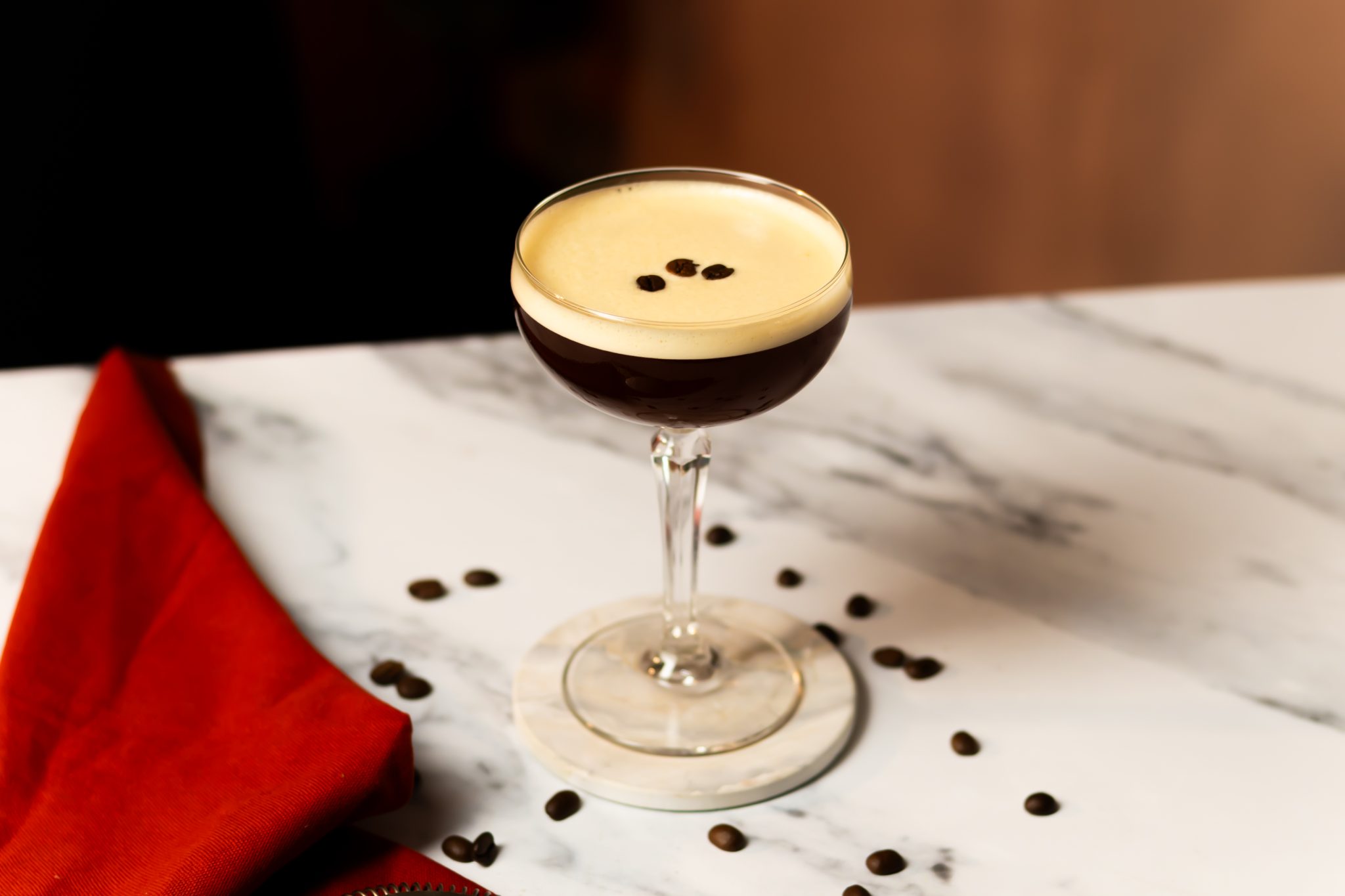 A side shot of a Mezcal Espresso Martini in a cocktail glass on a white coaster placed on a white marmol table surrounded by a red cloth and cofee beans.