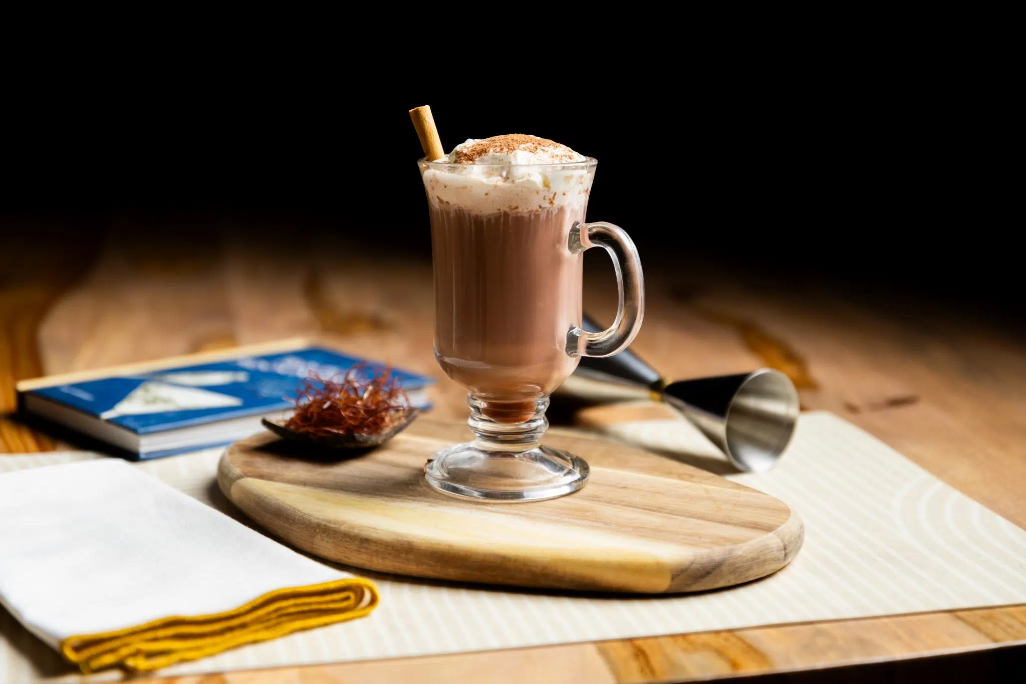 A side shot of a Mezcal Hot Chocolate cocktail in a mug on a wooden coardd on a wooden table surrounded by a white cloth, a jigger, a book and a small plate with chocolate shavings