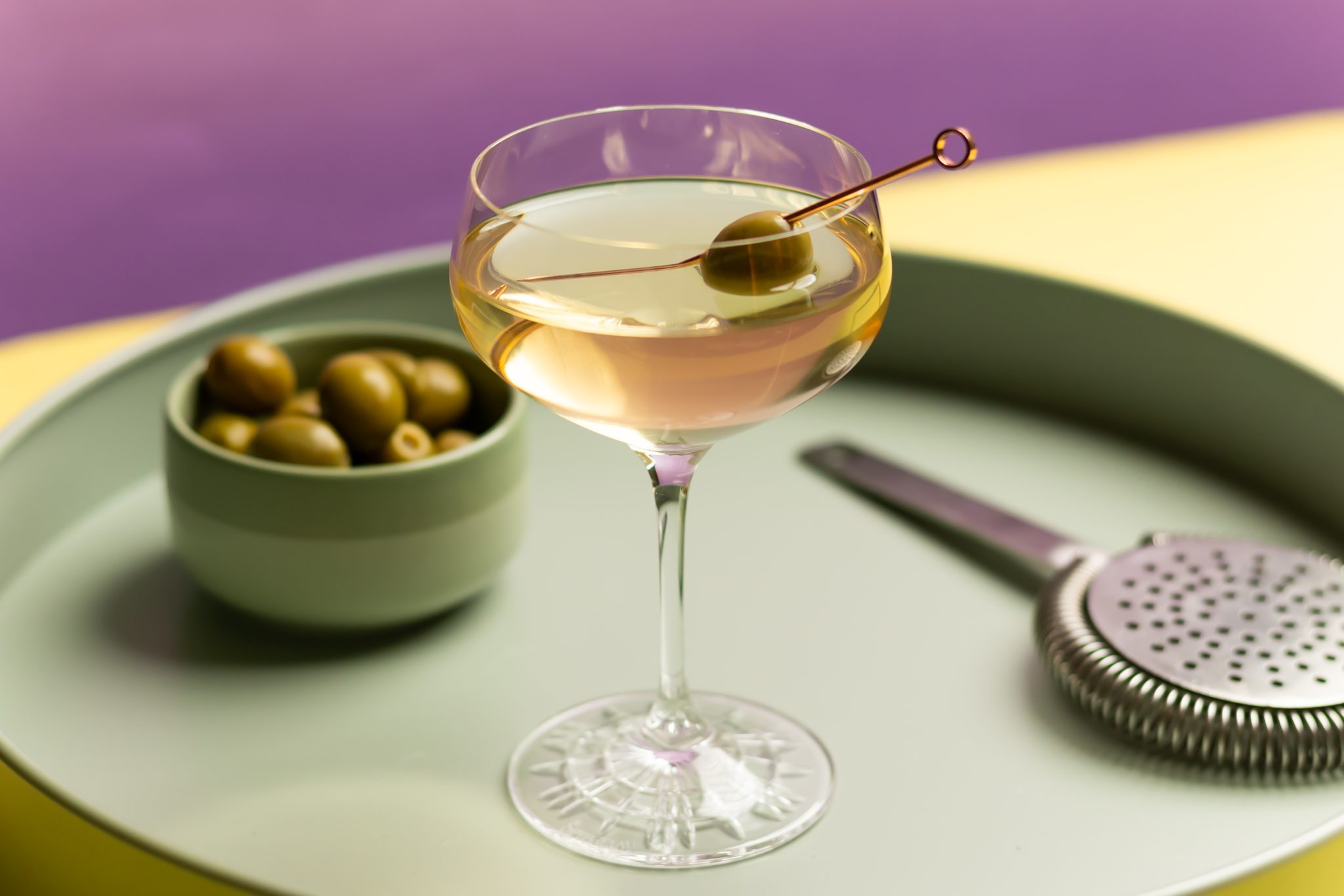 A side shot of a Mezcal Martini cocktail in a martini glass on a turquoise tray surrounded by a strainer and a bowl with olives inside, in front of a purple background