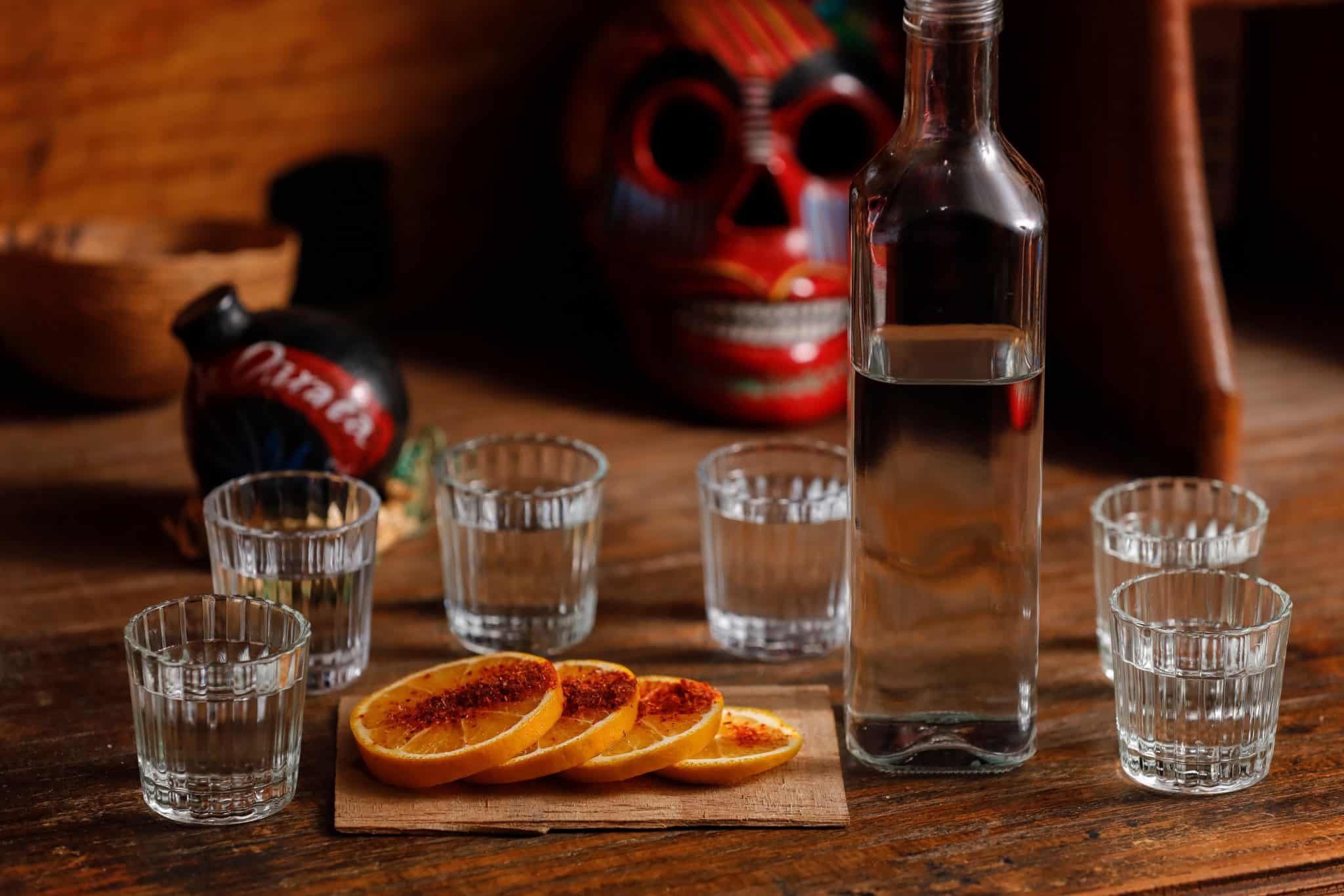 A bottle of mezcal with six shot glasses and four spiced orange slices on a wooden board.