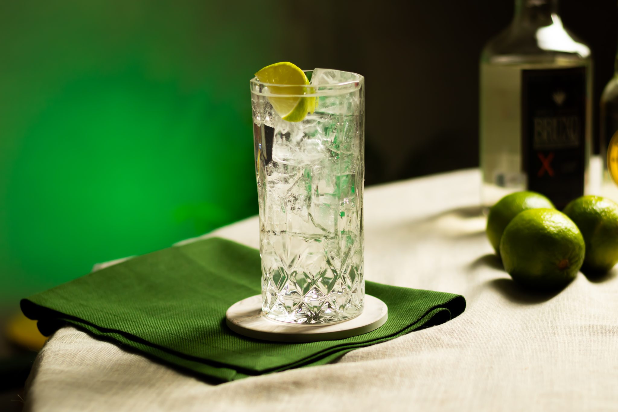 A side shot of a Mezcal and Tonic cocktail in a highball glass on a white coaster placed on a white table surrounded by three limes, a green cloth, and a mezcal bottle, in front of a green background