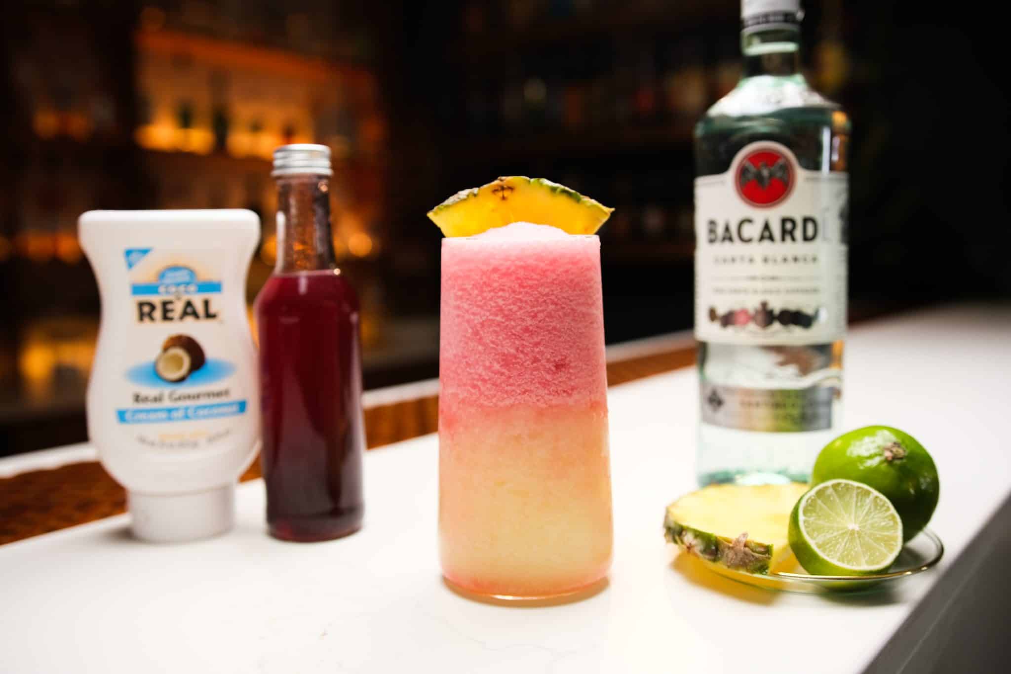 White rum, strawberry syrup, coconut syrup, lime and pineapple slice on a white bar table.