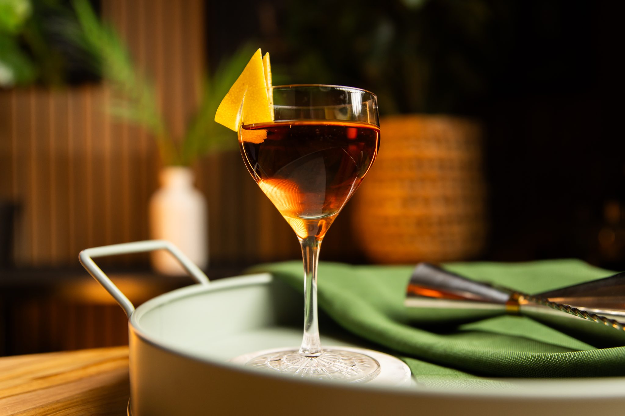 A side shot of a Midnight Bouquet cocktail in a coupe glass on a white coaster placed on a turquoise tray with a jigger and a green cloth on the side