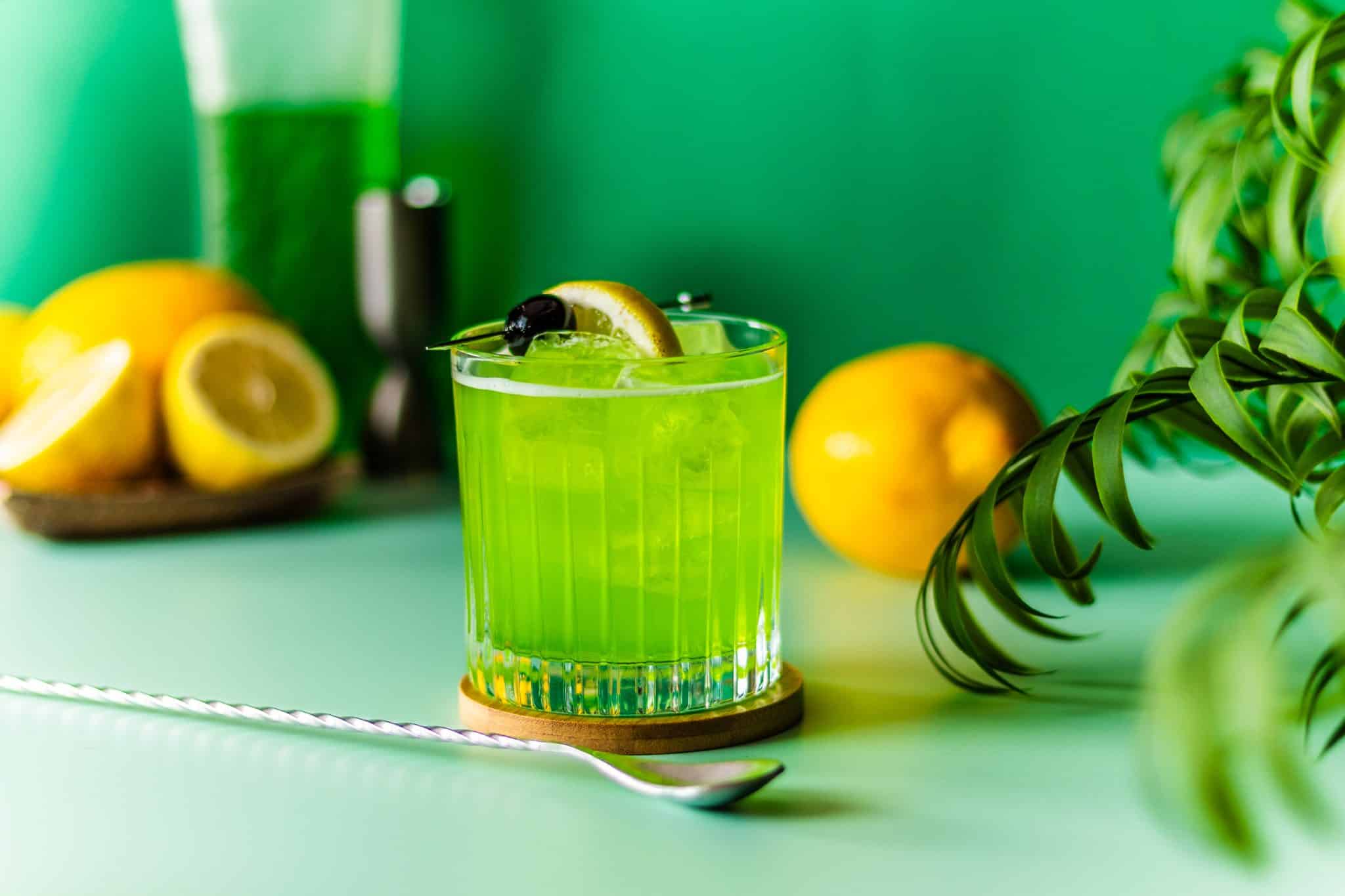 A side shot of a Midori Sour cocktail in an old-fashioned glass on a brown coaster placed on a green table with a bar spoon in front and some lemons on the background