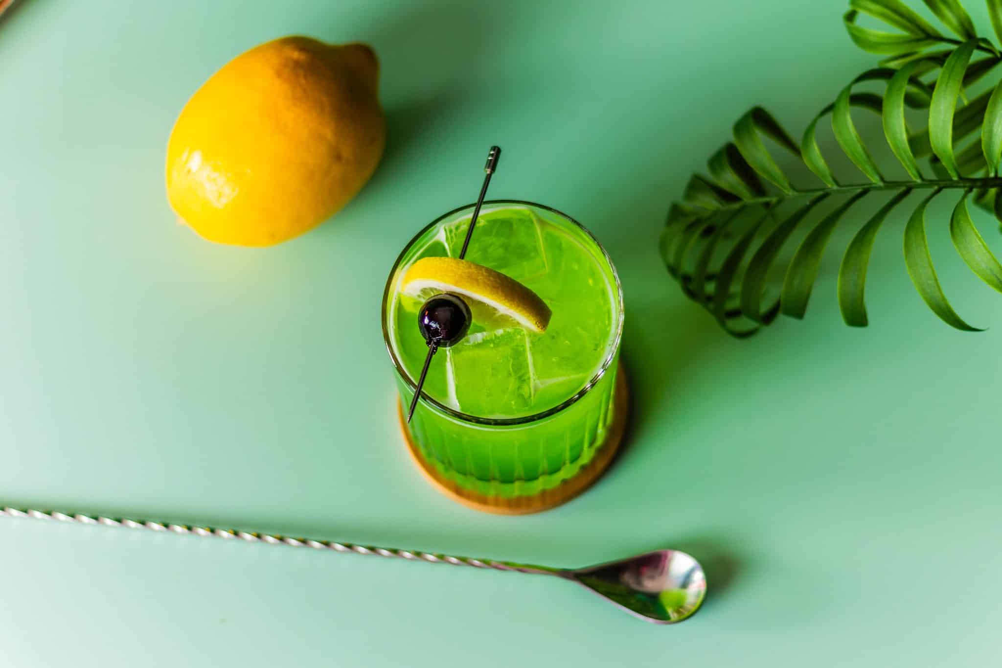 A Midori Sour cocktail, shot from above, in an old-fashioned glass on a brown coaster placed on a green table with a bar spoon in front and a lemon behind