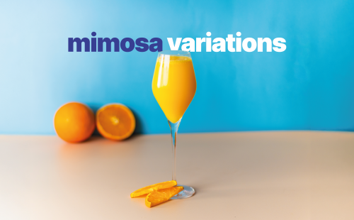 A shot of a mimosa cocktail in a wine glass on a light brown surface with two orange slice on the foot and two oranges behind on a blue background