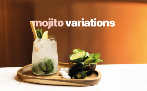 A shot of a type of mojito in a highball glass on a wooden tray with five sugar cubes, two japaleños and a wooden bowl with mint leaves and limes, all of that placed on a white surface in front of a orange background