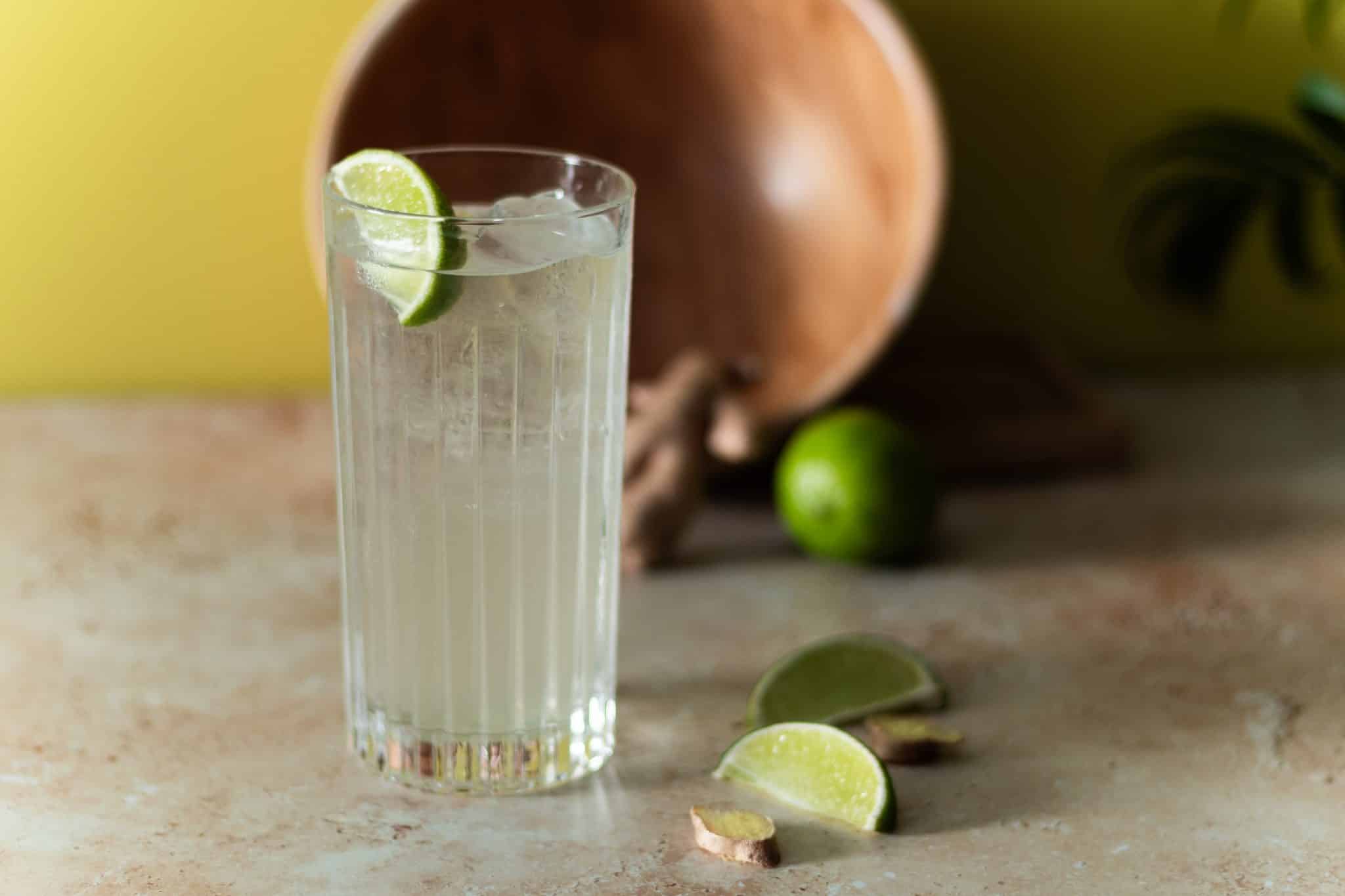 A side shot of a Moscow Mule cocktail in a highball glass on a grey-beige table with lemon wedges and ginger pieces on the side, and a brown wooden bowl on the background