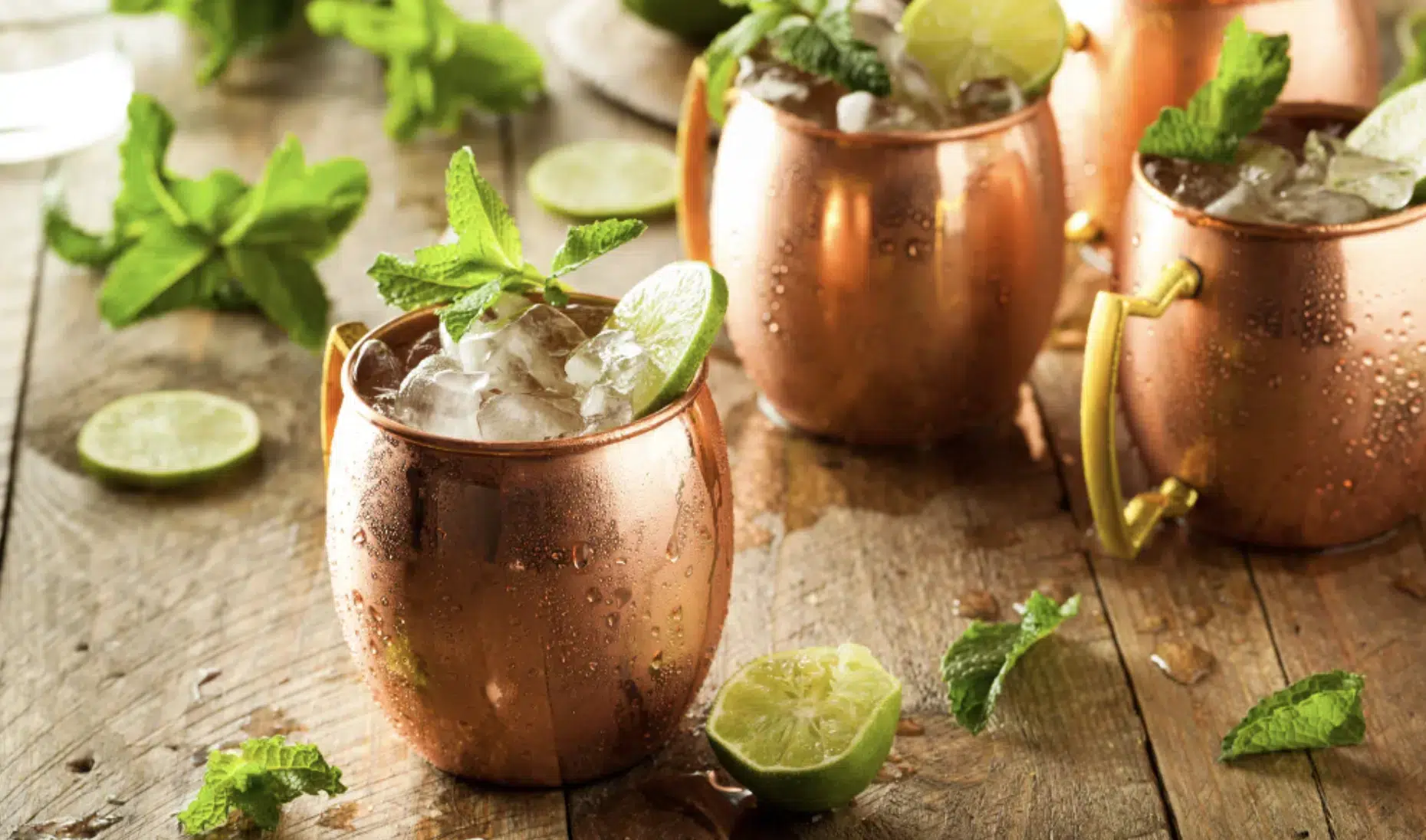 A shot of two Moscow Mule cocktail in copper mugs on a wooden surface surrounded by mint leaves and sprigs and lime slices