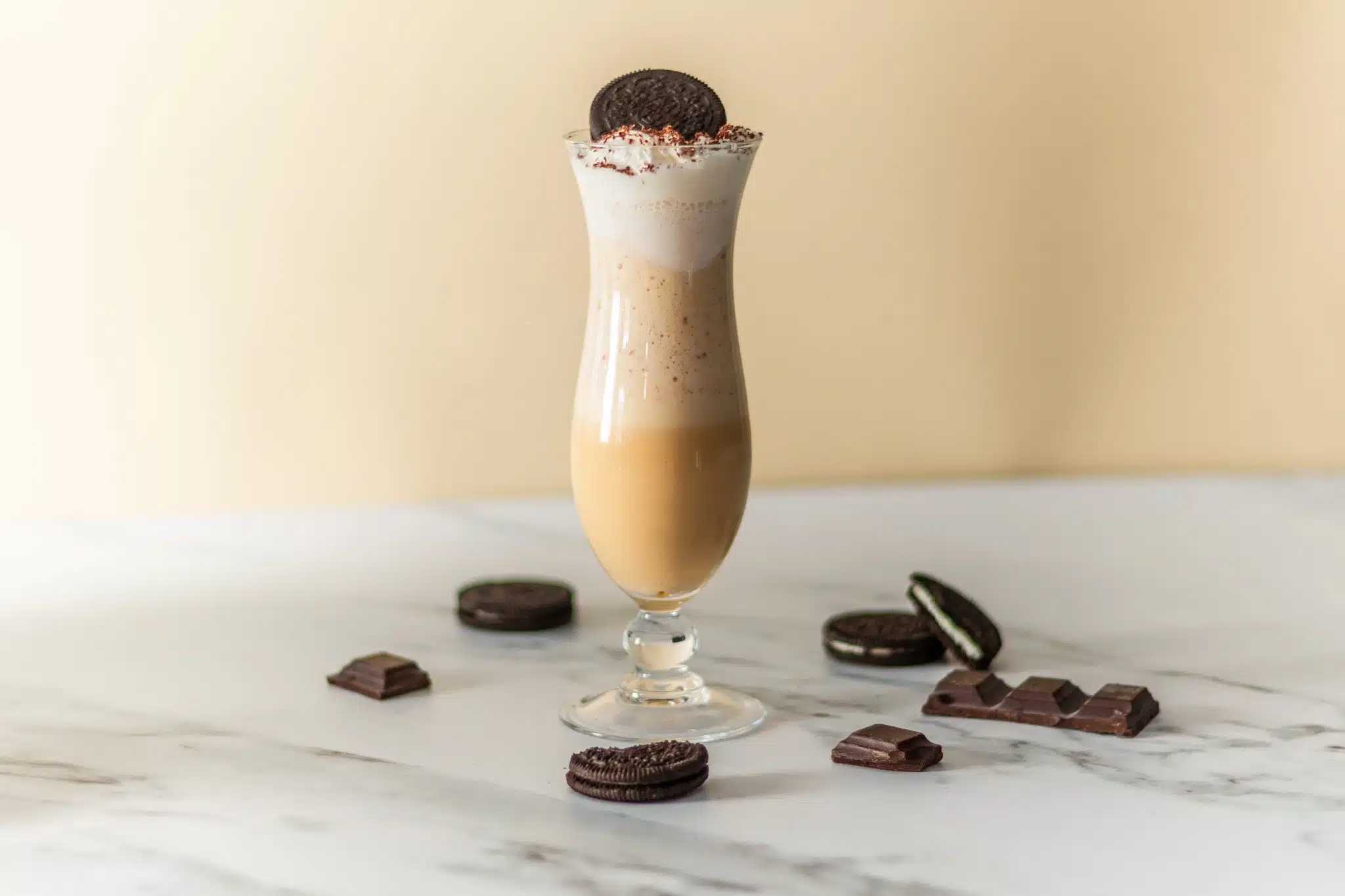 A side shot of a Mudslide cocktail in a hurricane glass on a white marmol table surrounded by Oreo cookies and chocolate blocks