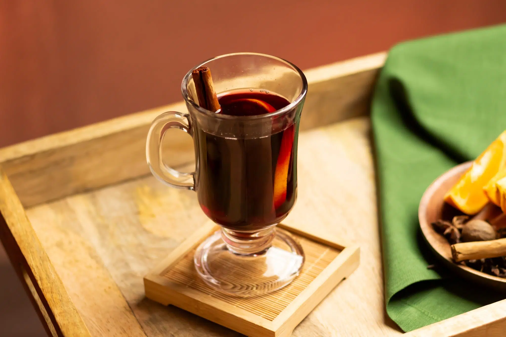A side shot of a Mulled Wine cocktail in a mug on a wooden coaster placed on a wooden tray surrounded bya green cloth and a plate with orange wedges, cinnamon stick, and star anise