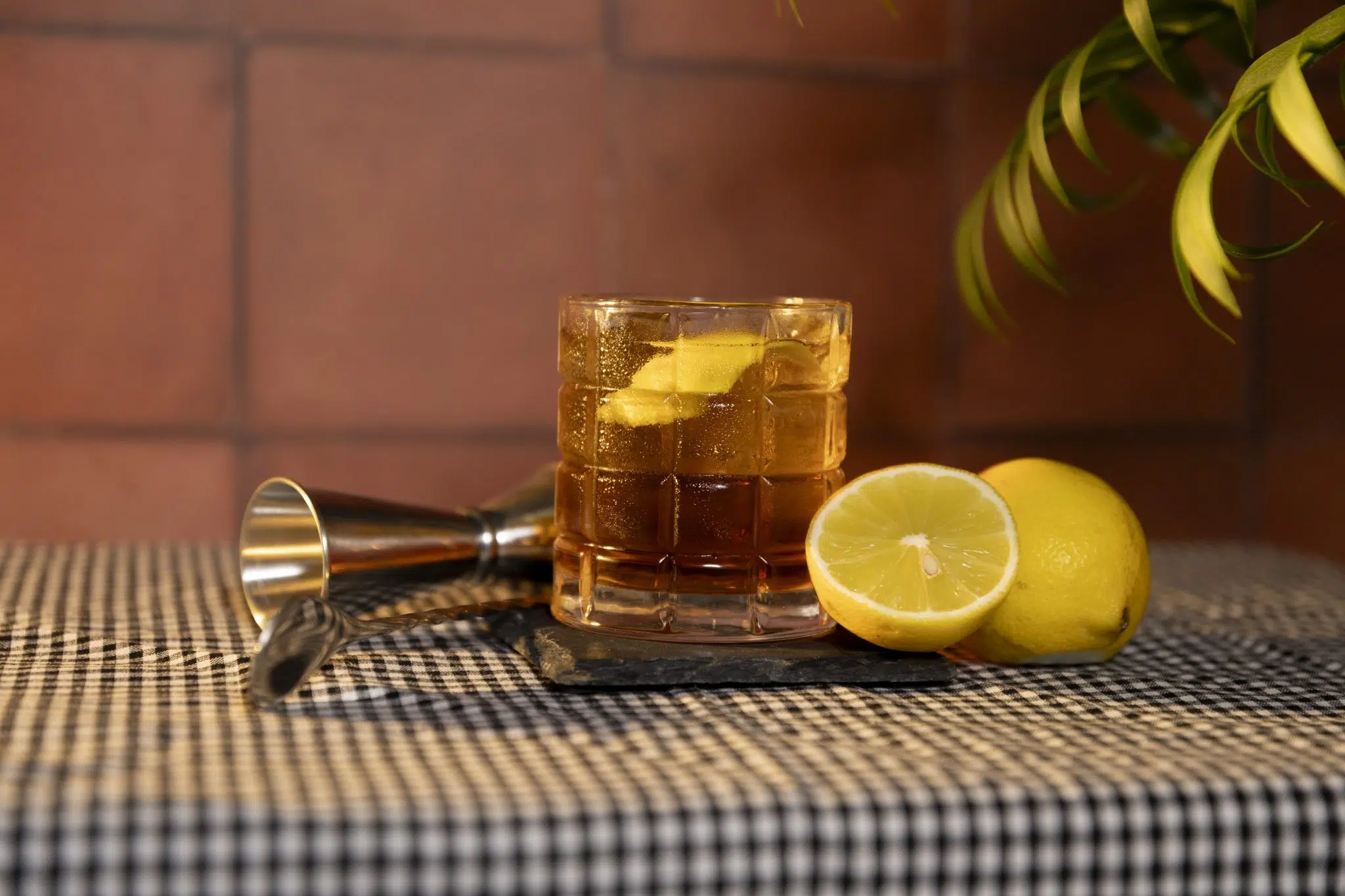 A side shot of a Nail In The Coffin cocktail in an old fashioned glass on a black stone coaster placed on a white and black checkered tablecloth surrounded by a jigger, a bar spoon, and two lemons