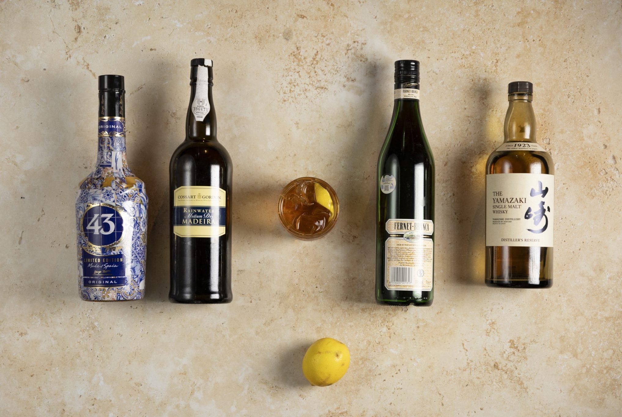 A Nail In The Coffin cocktail, shot from above, in an old fashioned glass on a beige surface surrounded by Japanese whisky, madeira, Licor 43, Fernet Branca and lemon