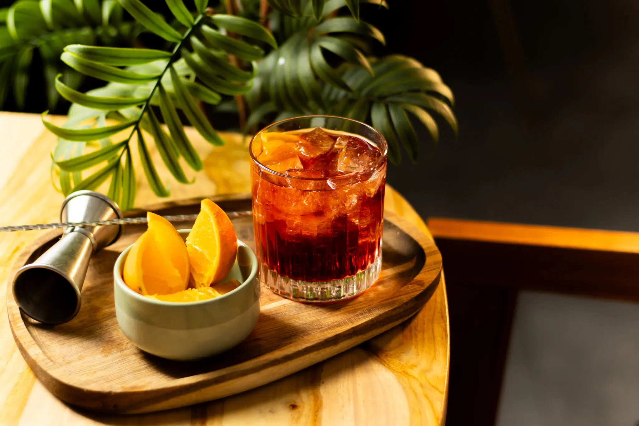 A side shot of a Negroni Sbagliato cocktail in an old fashioned glass on a wooden tray surrounded by a jigger, a bar spoon and a bowl with orange wedges.