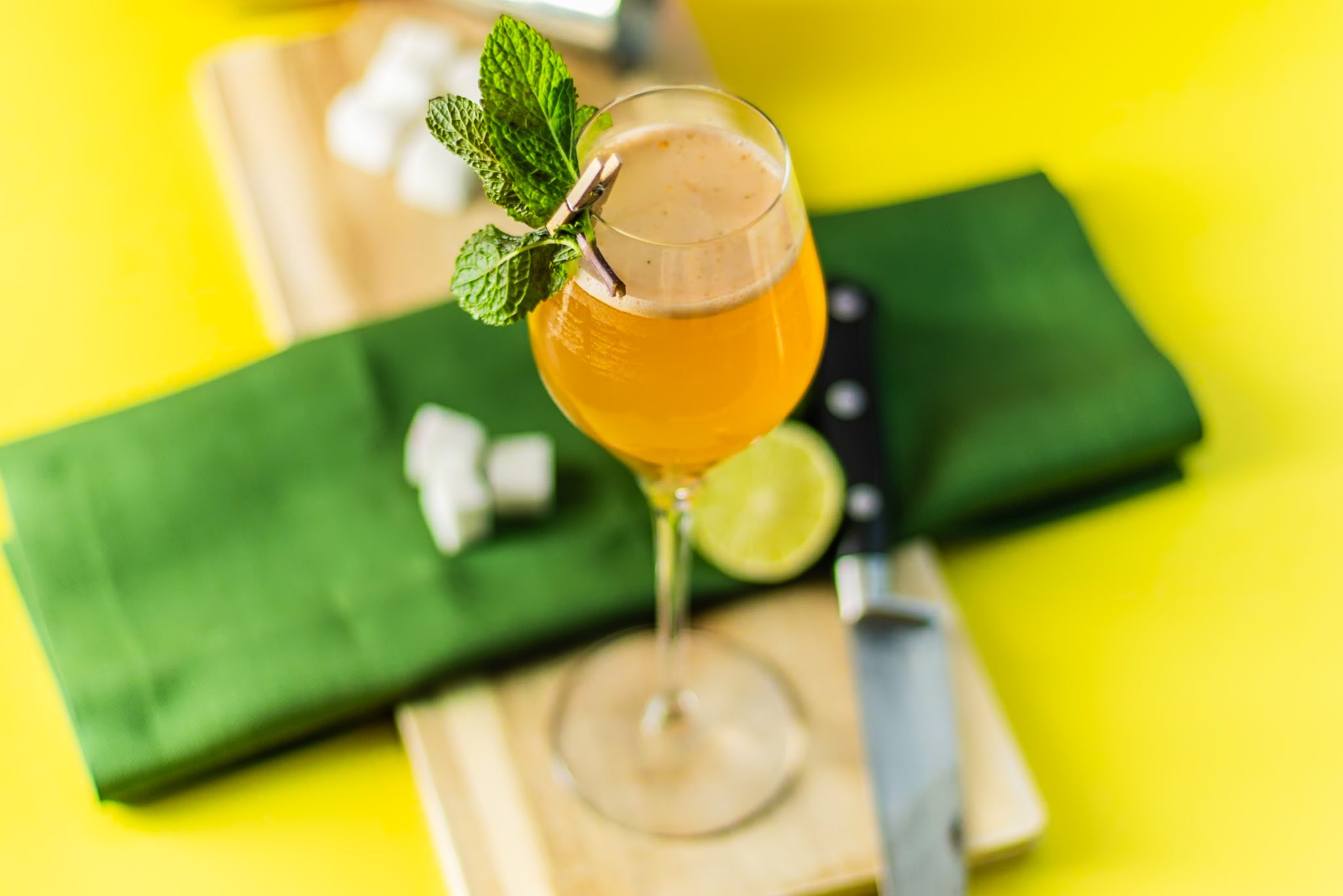 An Old Cuban cocktail, shot from above, in a campagne glass on a wooden board placed on a yellow table surrounded by a knife, a lime, sugar cubes, and a green cloth.