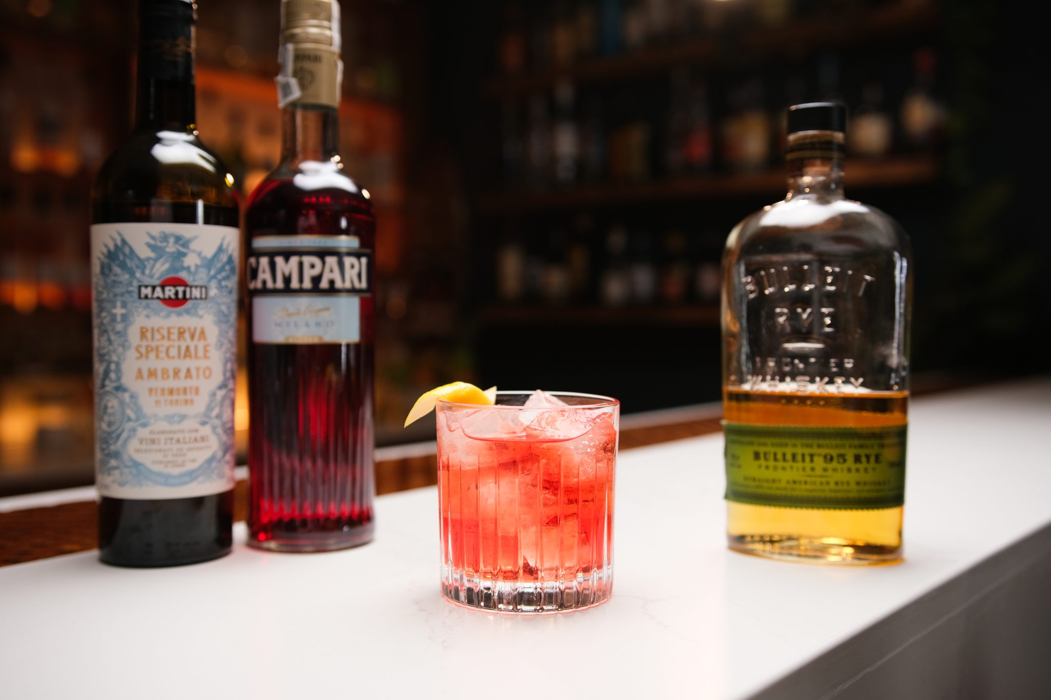 Rye Whiskey, Campari, and Dry Vermouth laid out on a white bar table.