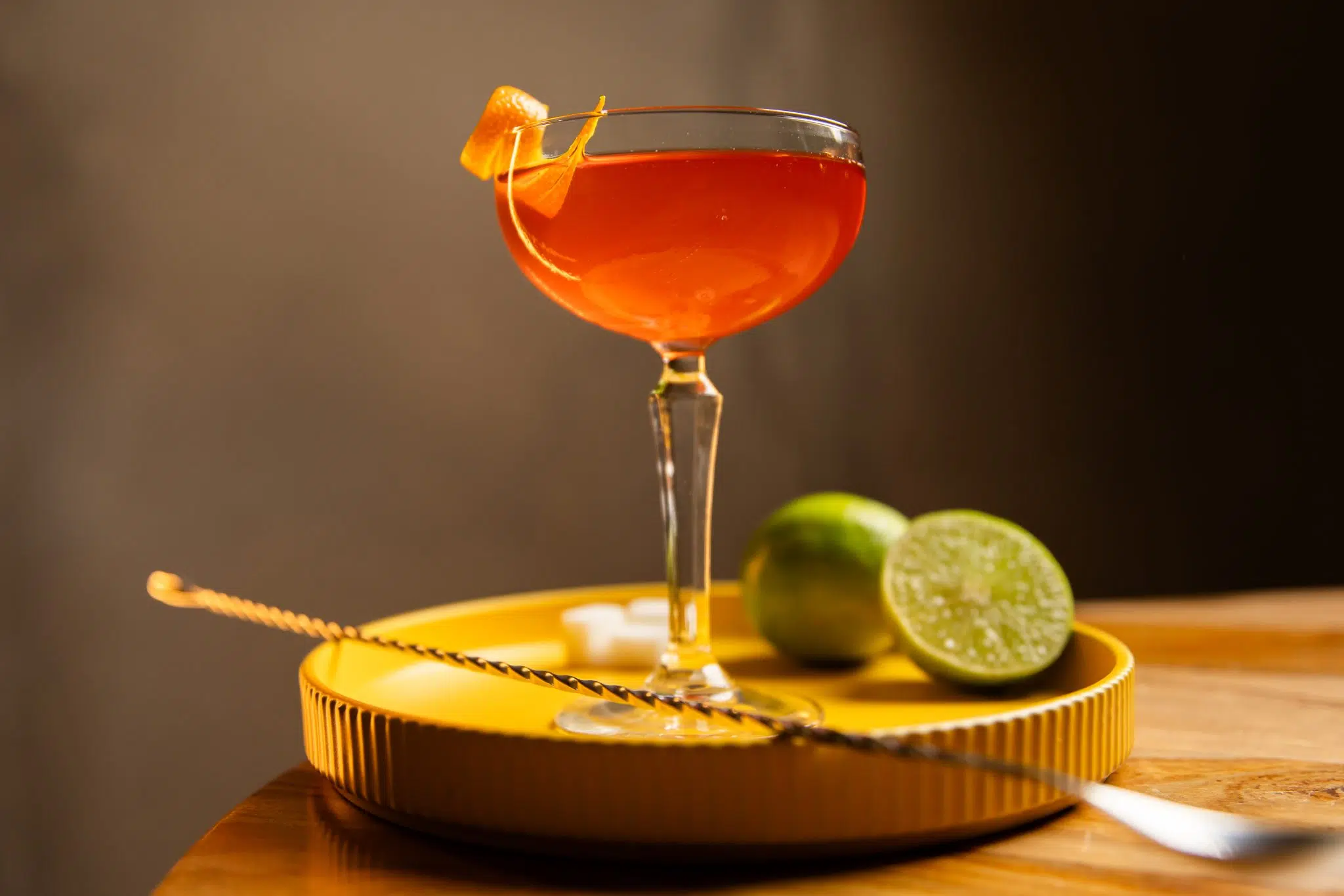 A side shot of an Oriental cocktail in a coupe glass on a yellow tray with two limes and a bar spoon around