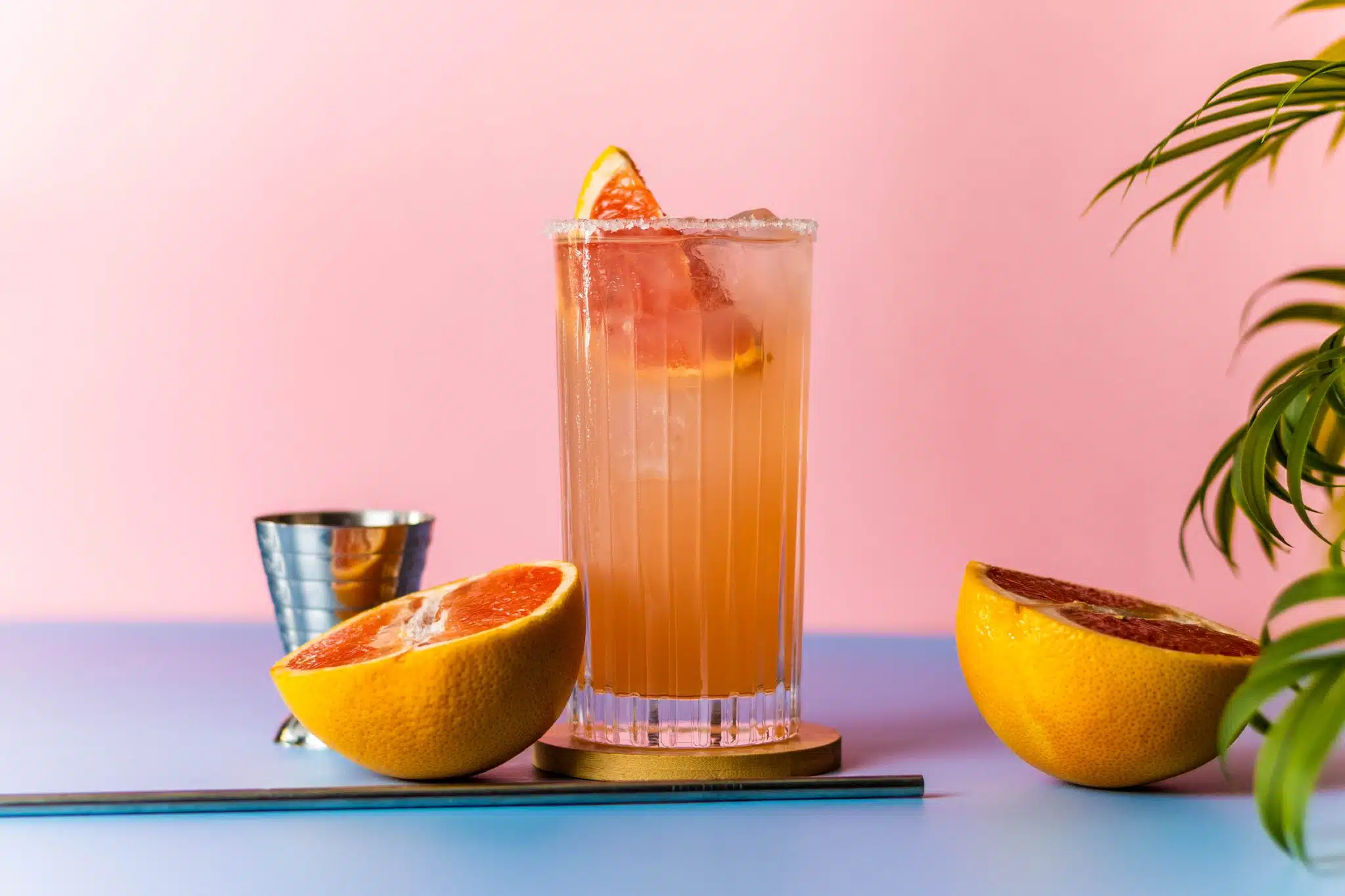 A side shot of a Paloma cocktail in a highball glass on a brown coaster placed on a light blue table with a jigger, a straw and two half grapefruits on the side