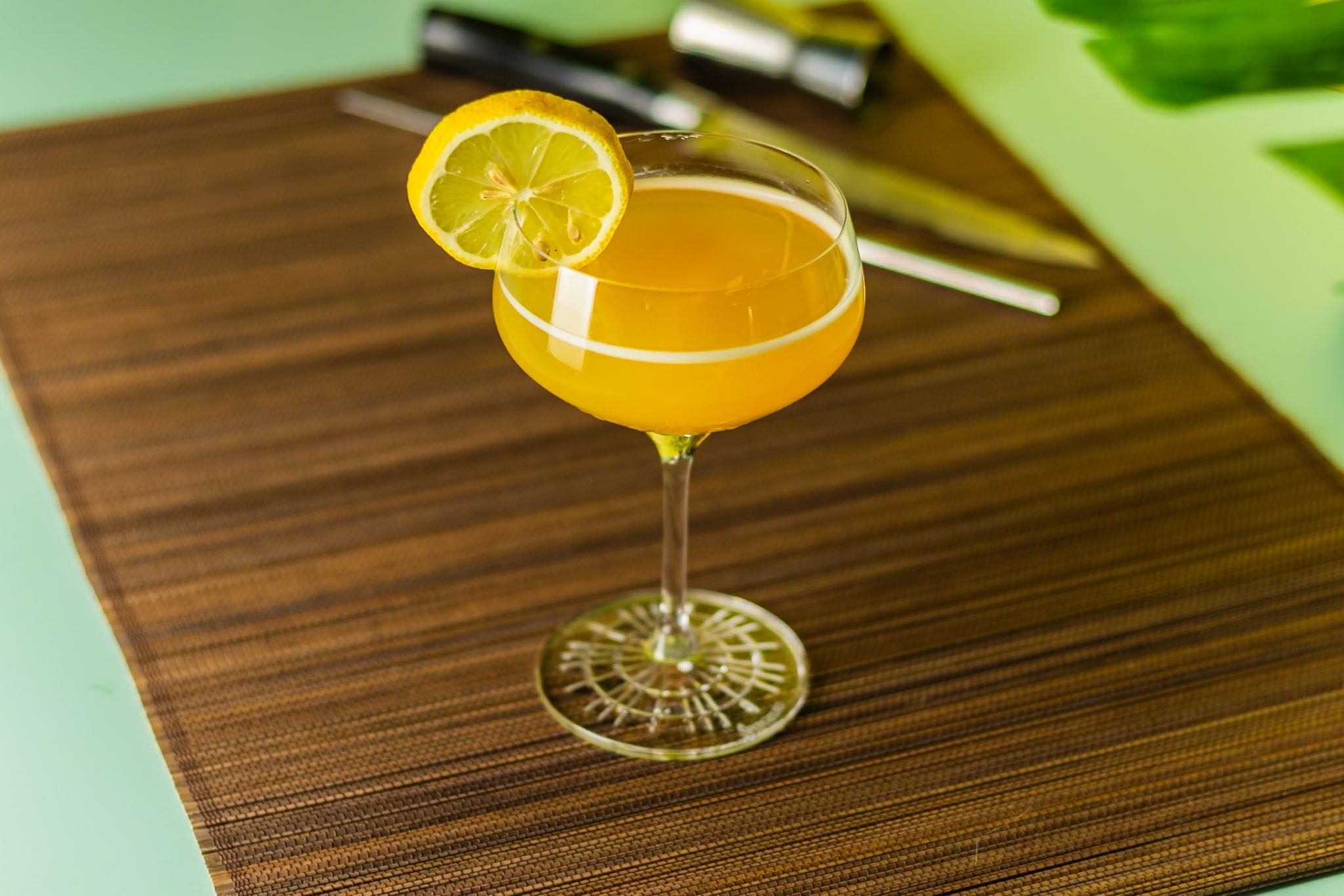 A Paradise cocktail, shot from above, in a cocktail glass on a dark brown placemat with a straw and a jigger around, placed on a turquoise table.
