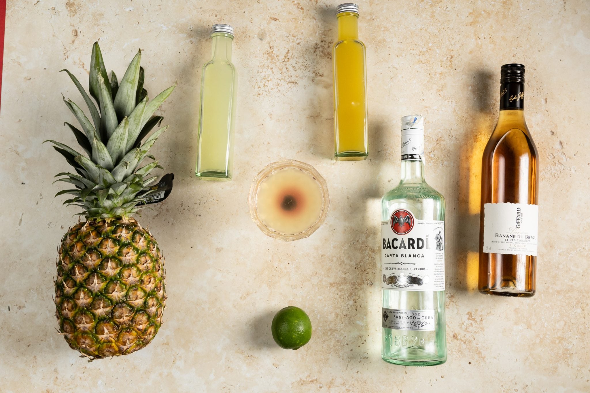 A Parasol Cocktail, shot from above, in a coupe glass on a beige surface surrounded by white rum, banana liqueur, pineapple juice, pineapple, lime juice, and lime