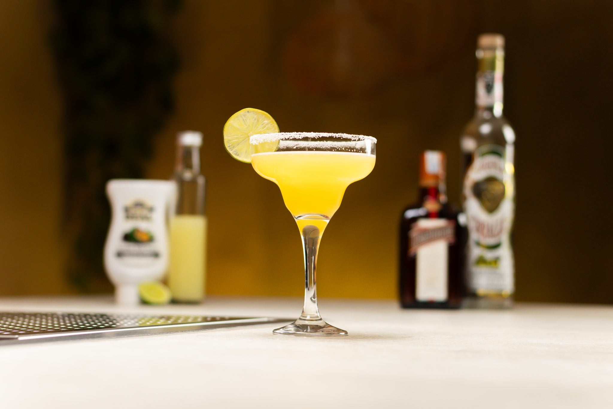 Tequila, orange liqueur, lime juice, lime, and passion fruit syrup laid out on a white bar table