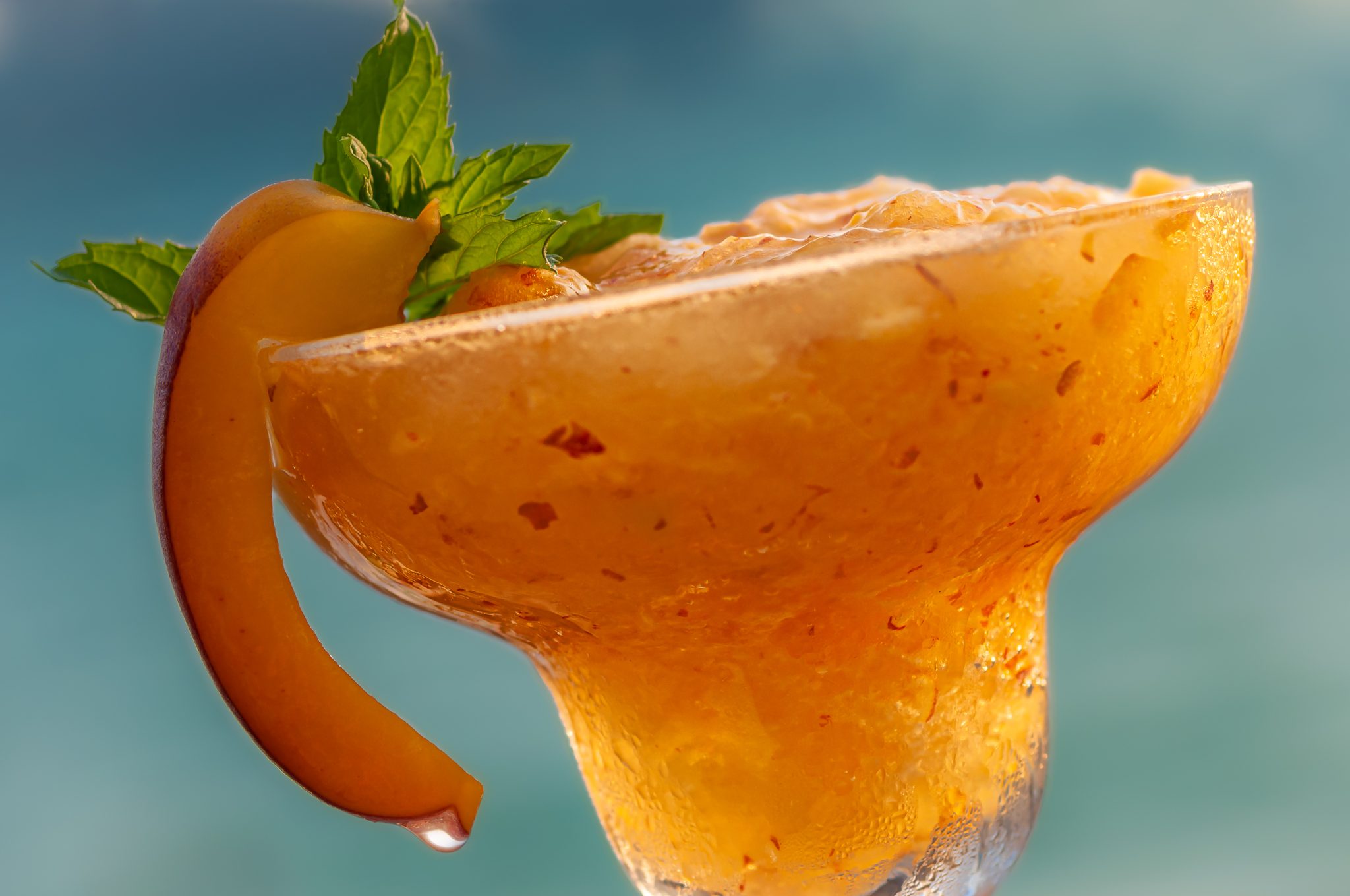 A Peach Margarita cocktail in a margarita glass garnish with a peach wedge, with a blurred blue background.