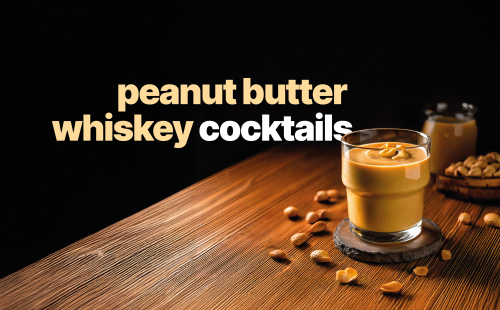 A side shot of a peanut butter whiskey cocktail in a low ball glass on a wooden coaster placed on a wooden surface surrounded by peanuts