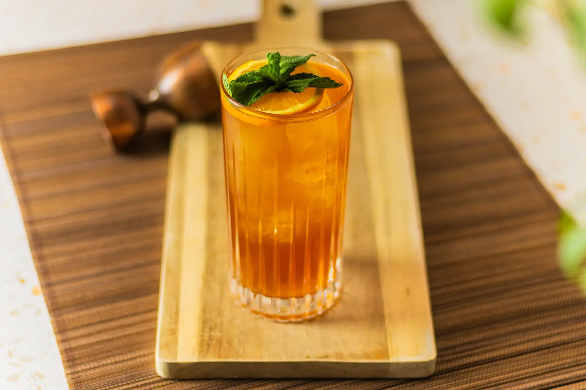 A side shot of a Pimm's Cup cocktail in a highball glass on a wooden board placed on a brown placemat with a jigger around.