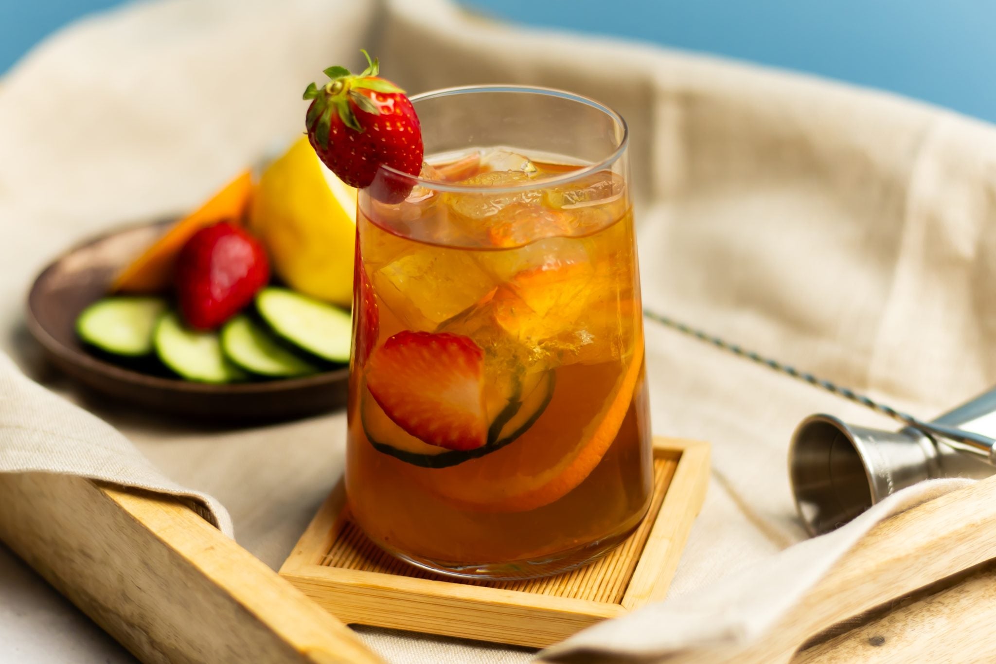 A side shot of a Pimm's Spritz cocktail in a wein glass on a wooden coaster placed on a wooden tray surrounded by a jigger, a bar spoon, a white cloth and a plate with fruit slices.
