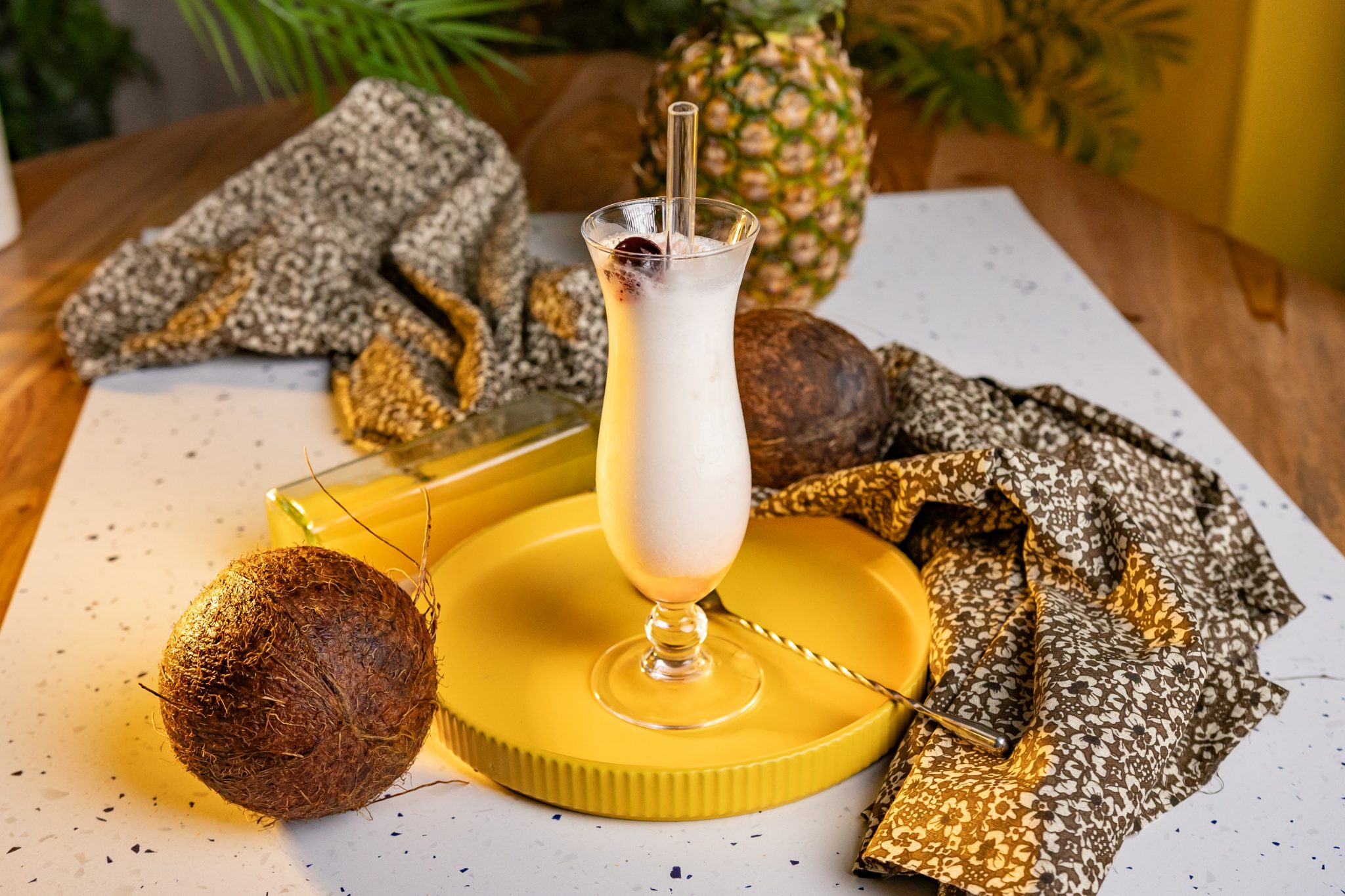 A side shot of a Piña Colada cocktail in a hurricane glass on a yellow tray surrounded by two coconuts, a pineapple, a bar spoon and a grey cloth