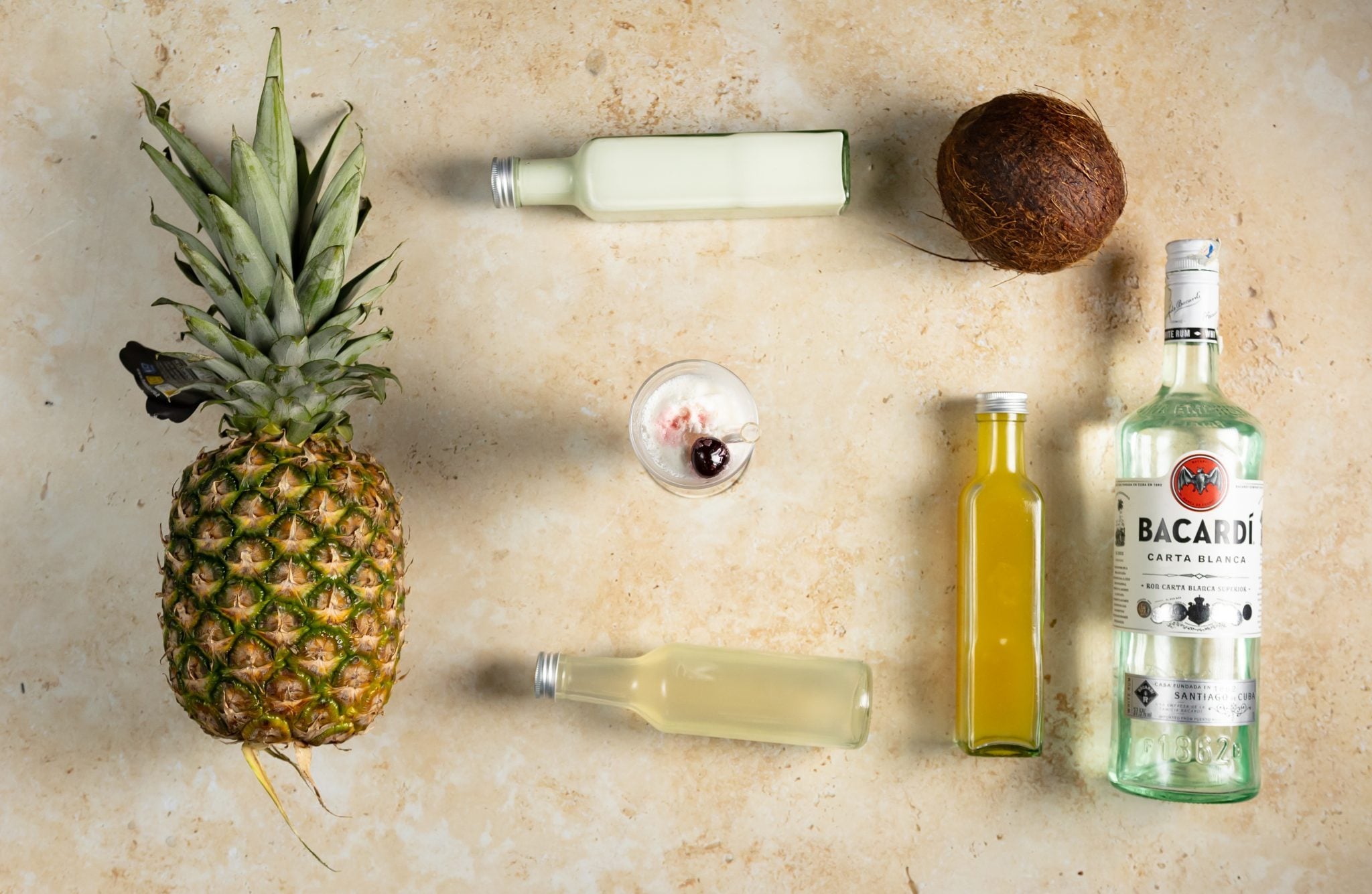 A Piña Colada cocktail, shot form above, in a hurricane glass on a beige surface surrounded by White Rum, coconut cream, coconut, pineapple, pineapple juice, and lime juice
