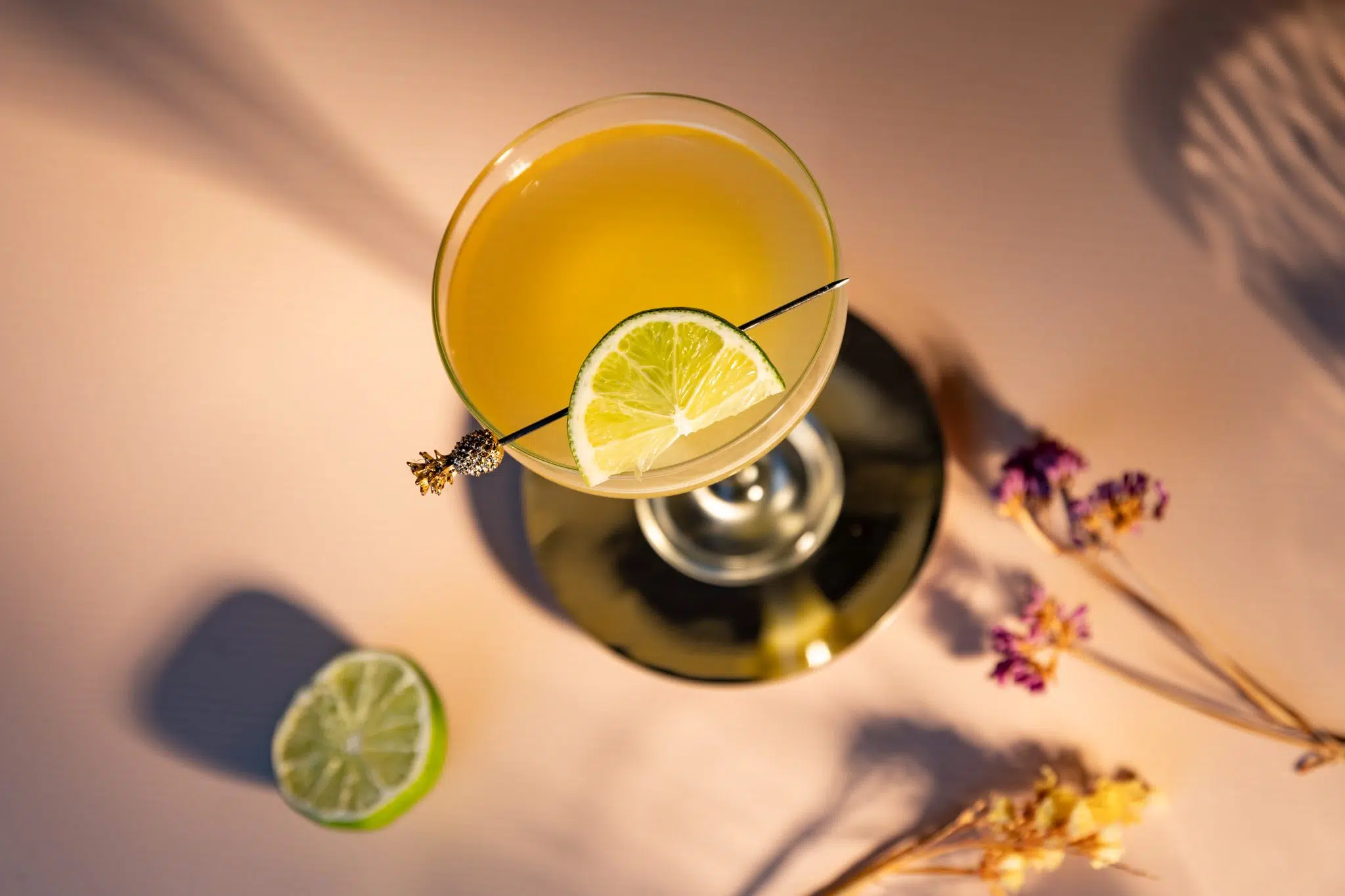 A shot from above of a Pineapple Margarita cocktail in a margarita glass on a plate placed on a white surface surrounded by a lime and two flower sprigs