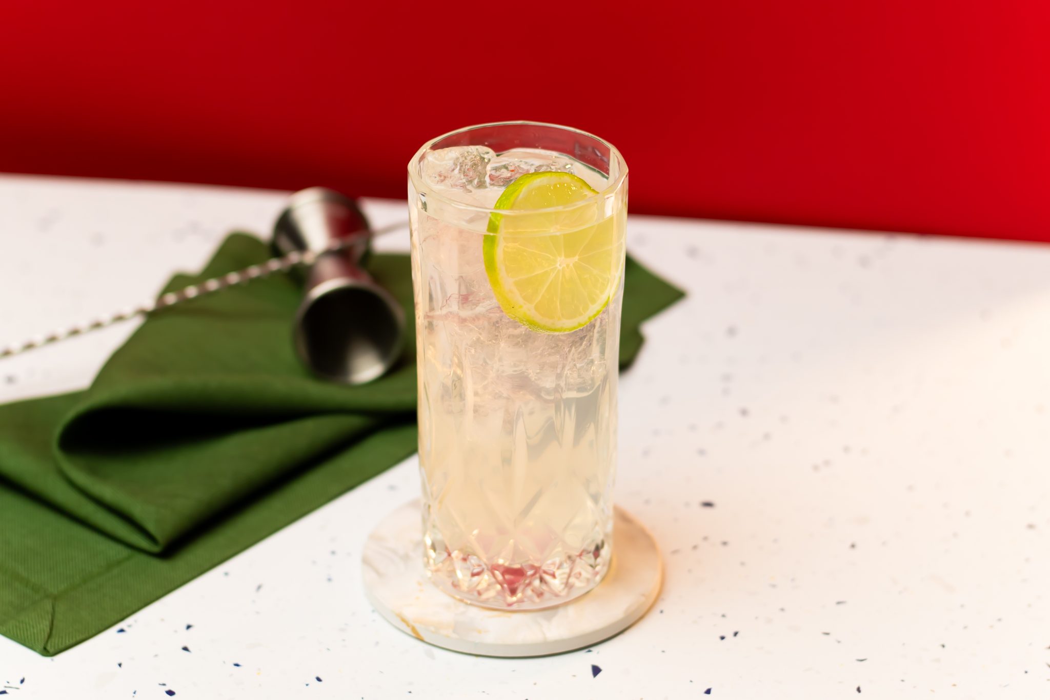 A side shot of a Pisco Collins cocktail in a highball glass on a white coaster placed on a white surface surrounded by a jigger, a bar spoon and a green cloth, in front of a red background.