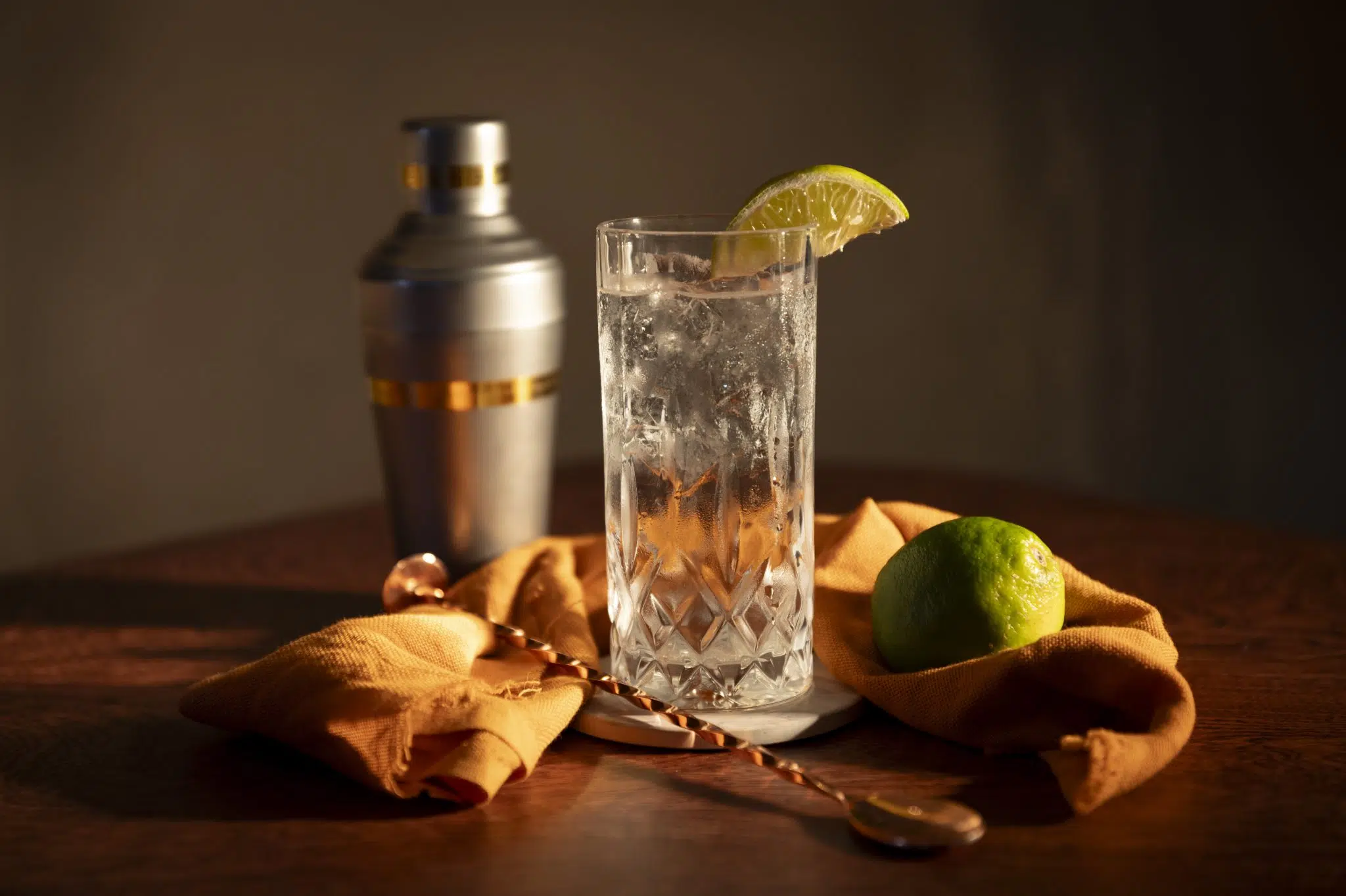 A side shot of a Pisco Highball cocktail in a highball glass on a wooden coaster placed on a brown surface surrounded by a lime, a bar spoon, a cocktail shaker and an orange cloth