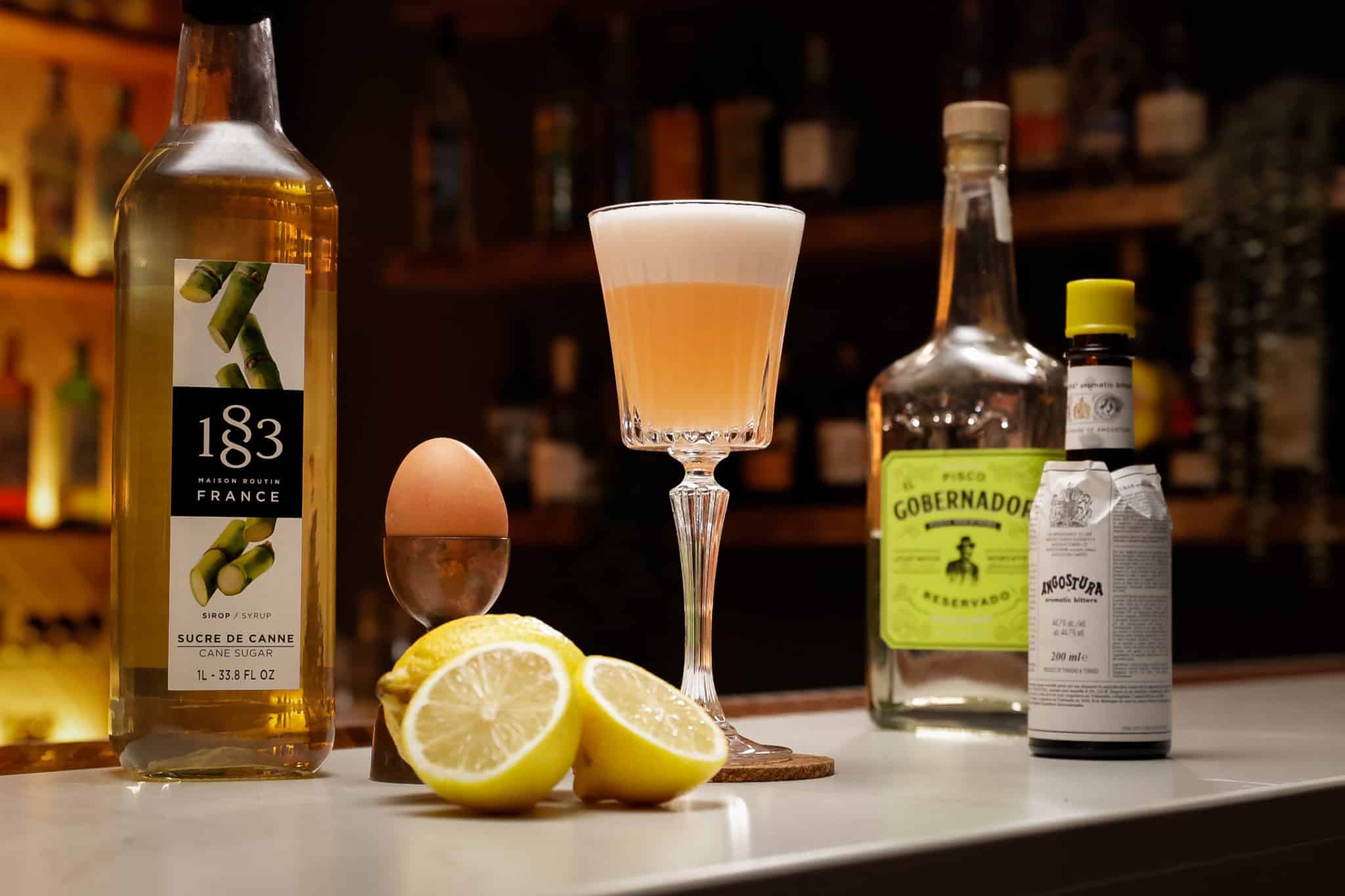 Pisco, Lime Juice, Simple Syrup, Egg and Angostura Bitters laid out on a white bar table