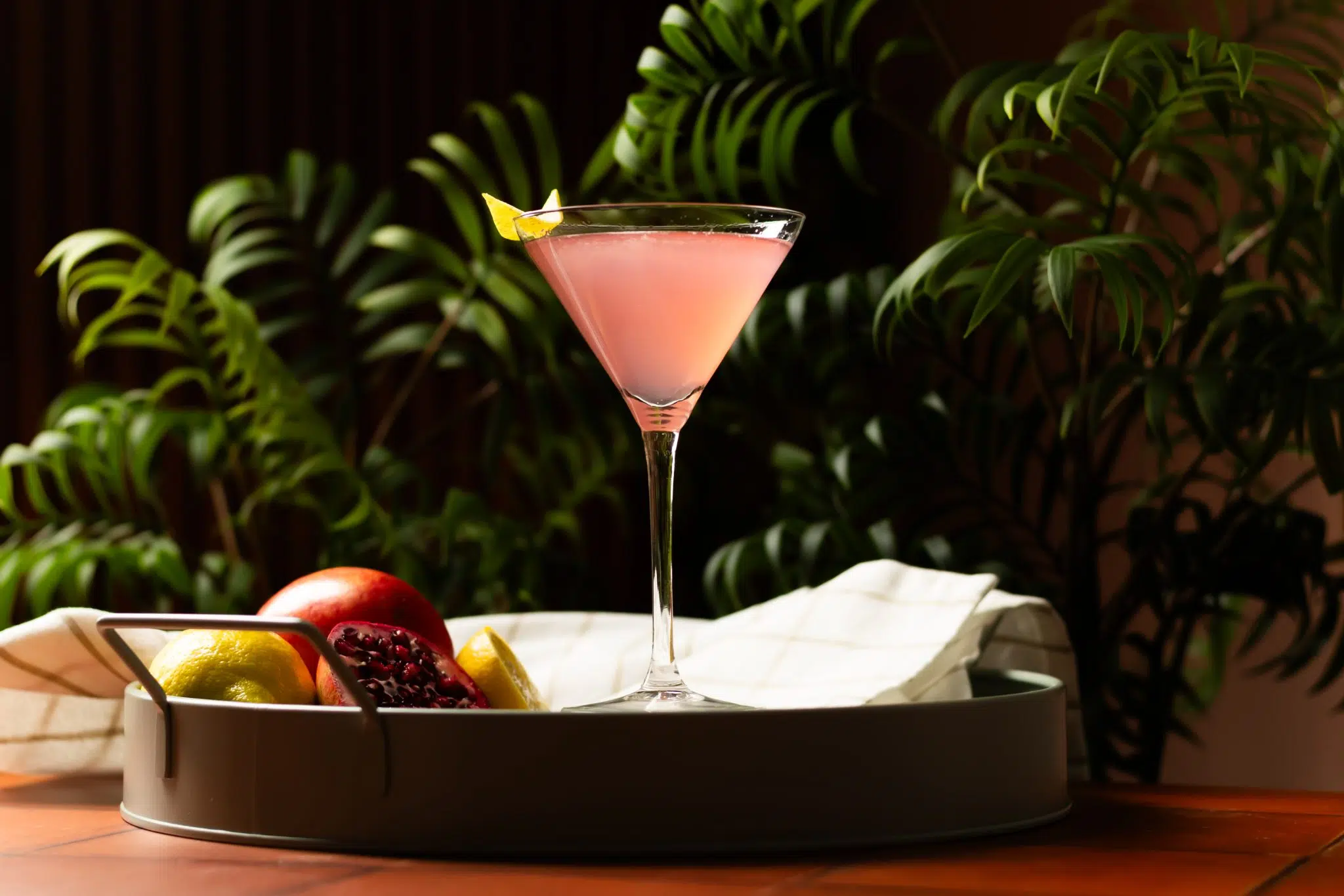 A side shot of a Pomegranate Martini cocktail in a martini glass on a yellow tray surrounded by two pomegranates, two lemons, and a white cloth