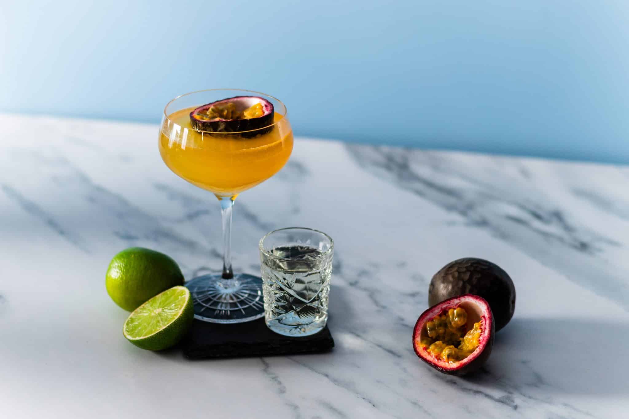 A side shot of a Pornstar Martini cocktail in a martini glass on a black stone coaster with a Prosecco shot, limes and passionfruit, placed on a white marmol table, in front of a blue wall.