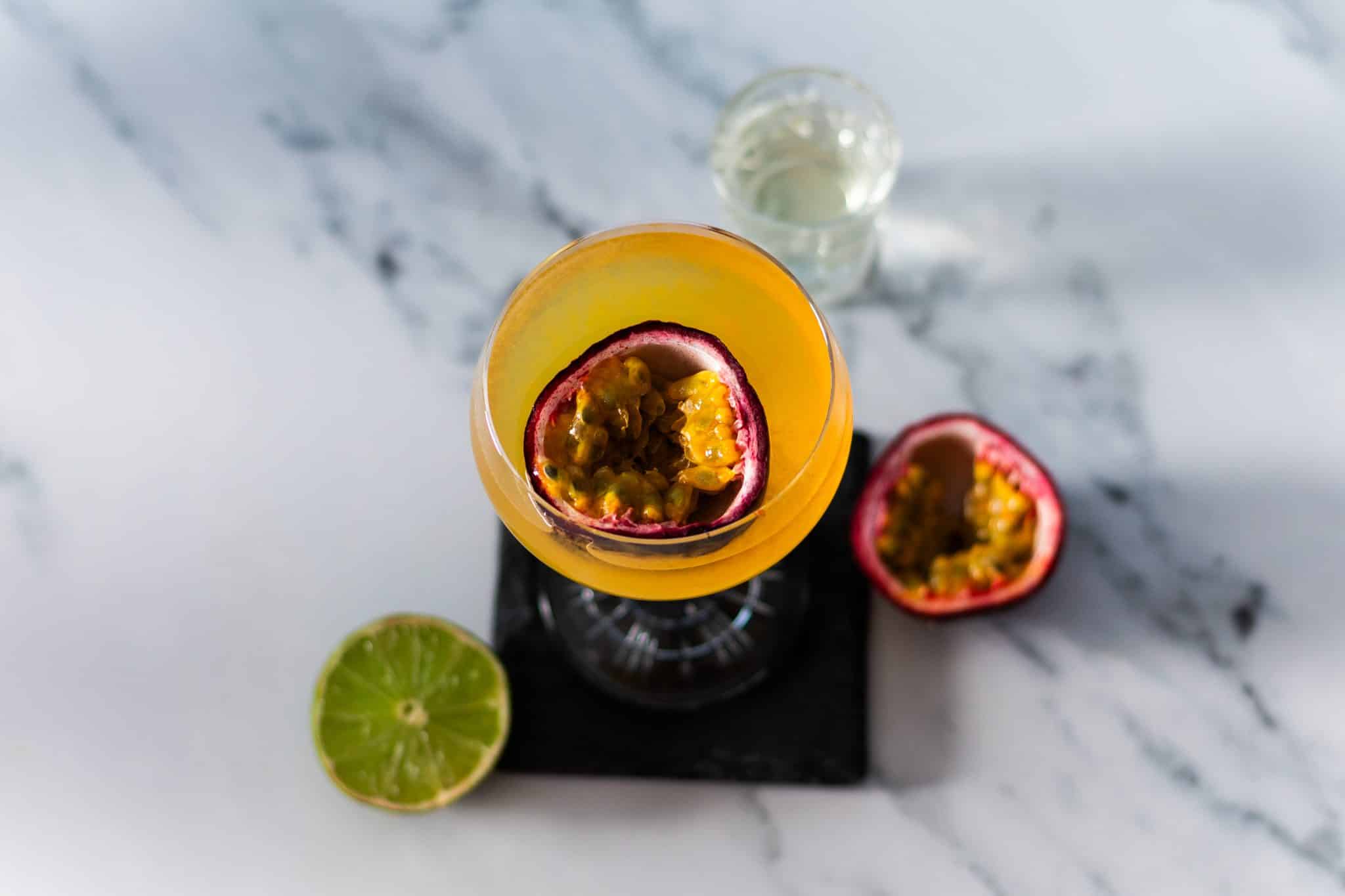 A Pornstar Martini cocktail, shot from above, in a martini glass on a black stone coaster with a Prosecco shot, half lime, and half passionfruit placed on a white marmol table.