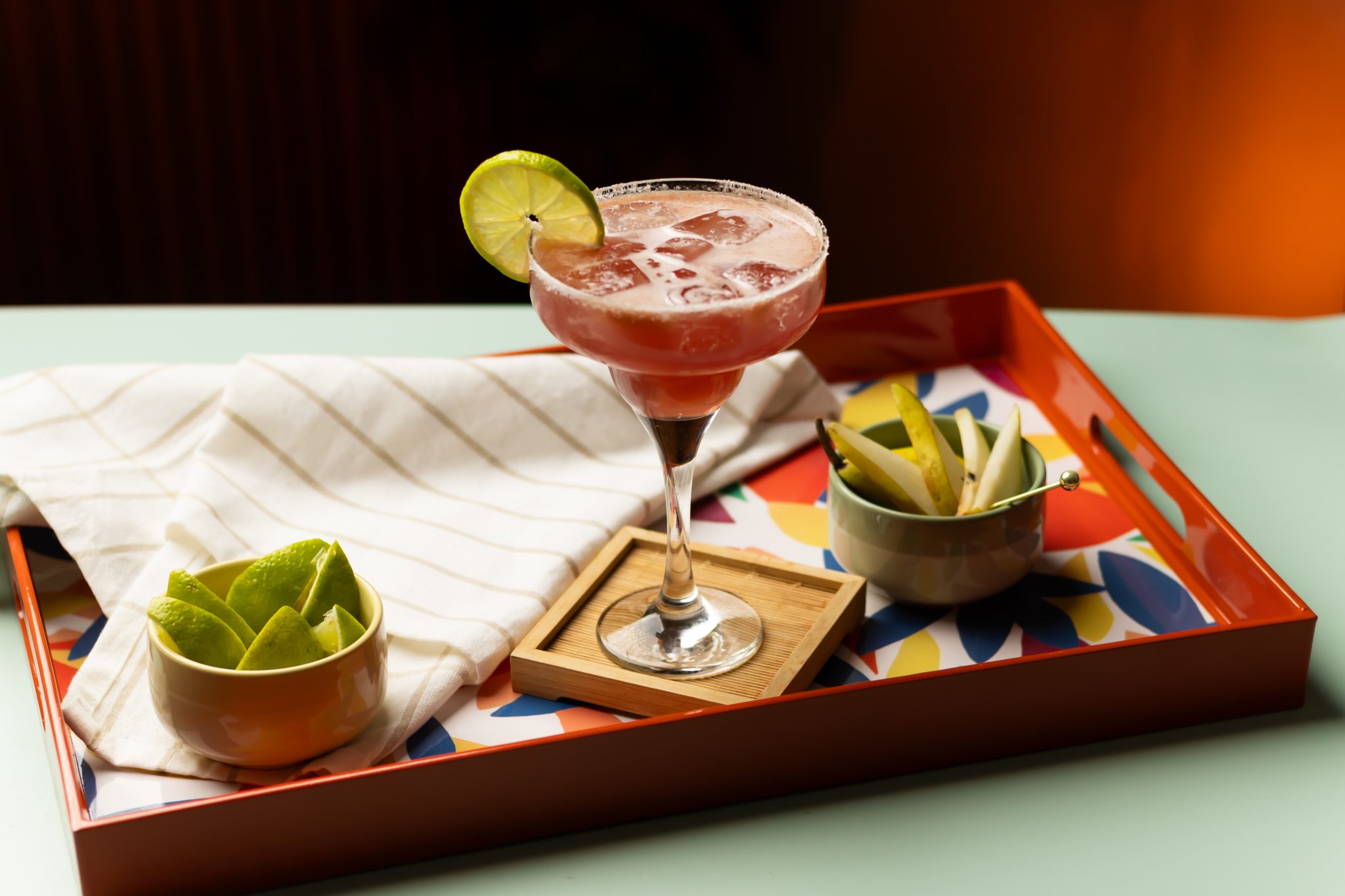A side shot of a Prickly Pear Margarita cocktail in a margarita glass on a wooden coaster placed on a red tray surrounded by a bowl with lime wedges, a bowl with apple slices and a white cloth