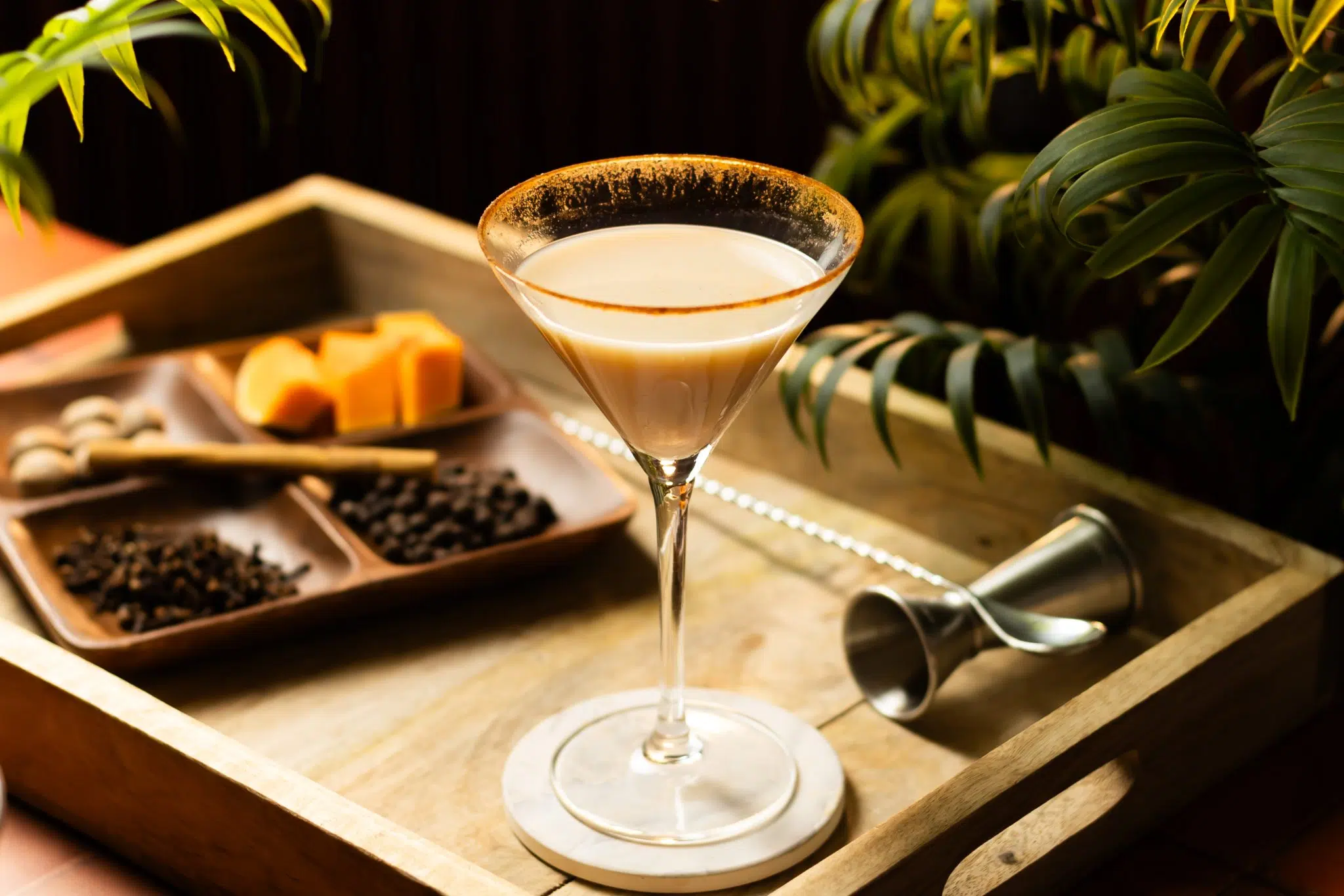 A side shot of a Pumpkin Spice Martini cocktail in a martini glass on a white coaster placed on a wooden tray surrounded by a jigger, a bar spoon, and a plate with different types of spices and pumpkin