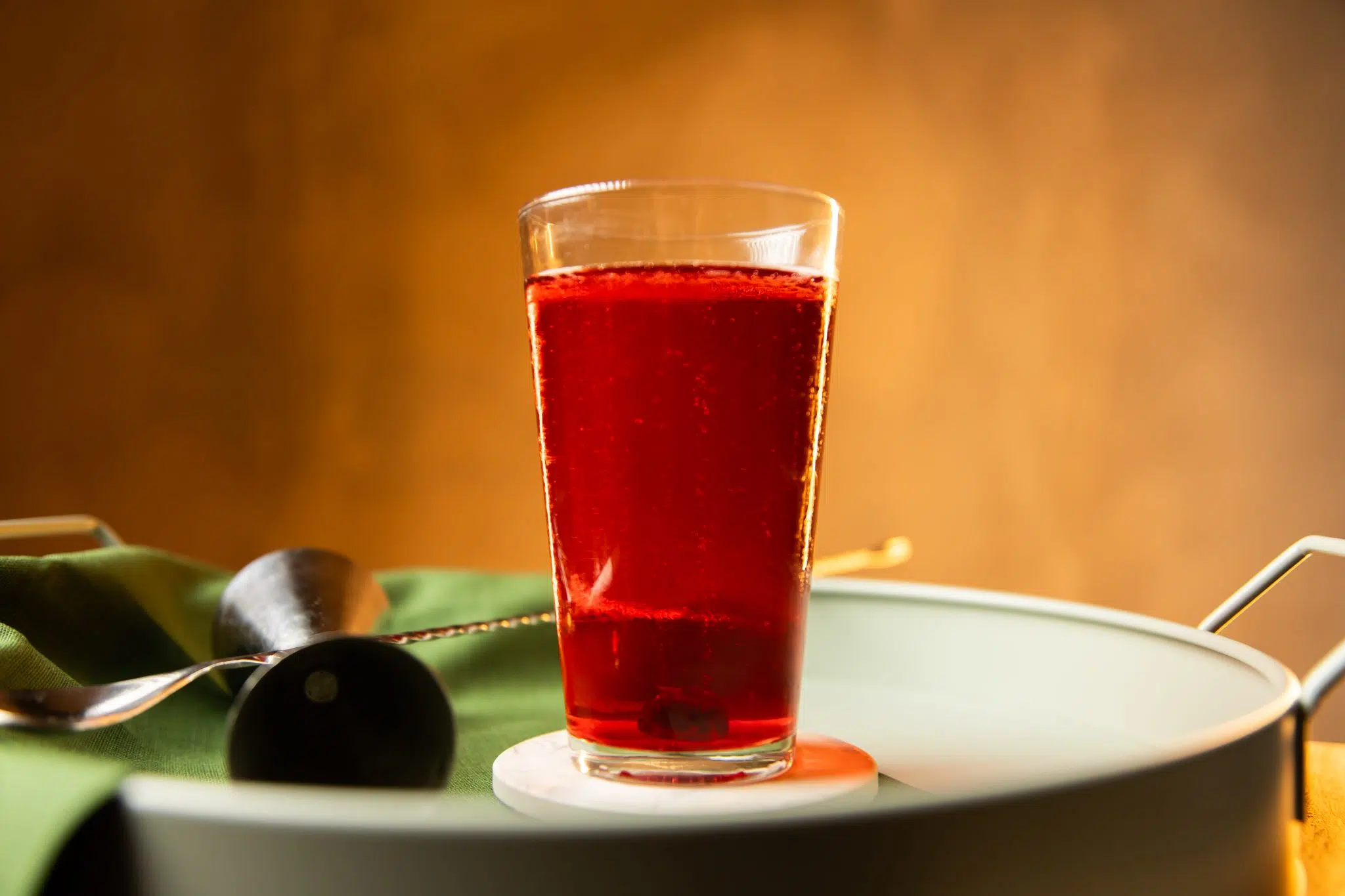 A side shot of a Queen Mary cocktail in a highball glass on a white coaster placed on a turquoise tray surrounded by a jigger, a bar spoon and a green cloth