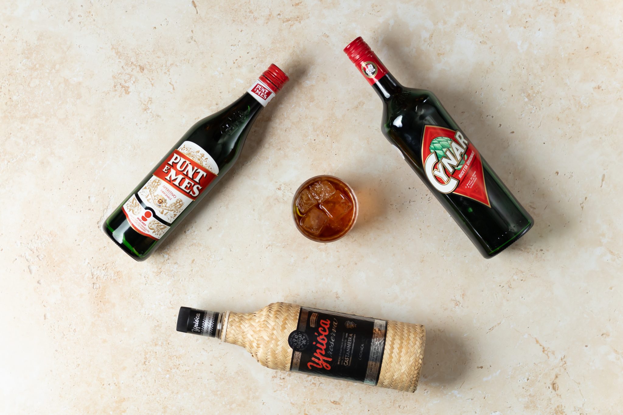 A Rabo de Galo cocktail, shot from above, in an old fashioned glass on a beige surface surrounded by Cachaça, red Vermouth, and Cynar