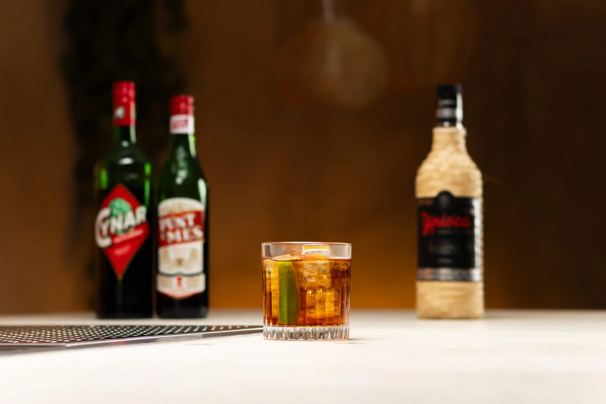 Cachaça, red Vermouth, and Cynar laid out on a white bar table