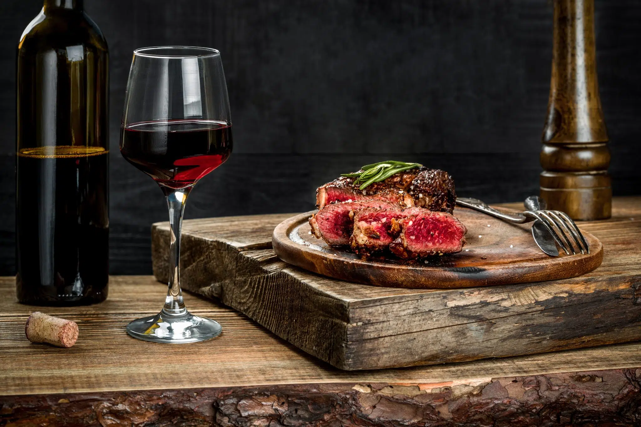 A side shot of a wooden table with a red wine glass, a red wine bottle and a plate with grilled beef meat cutted in slices